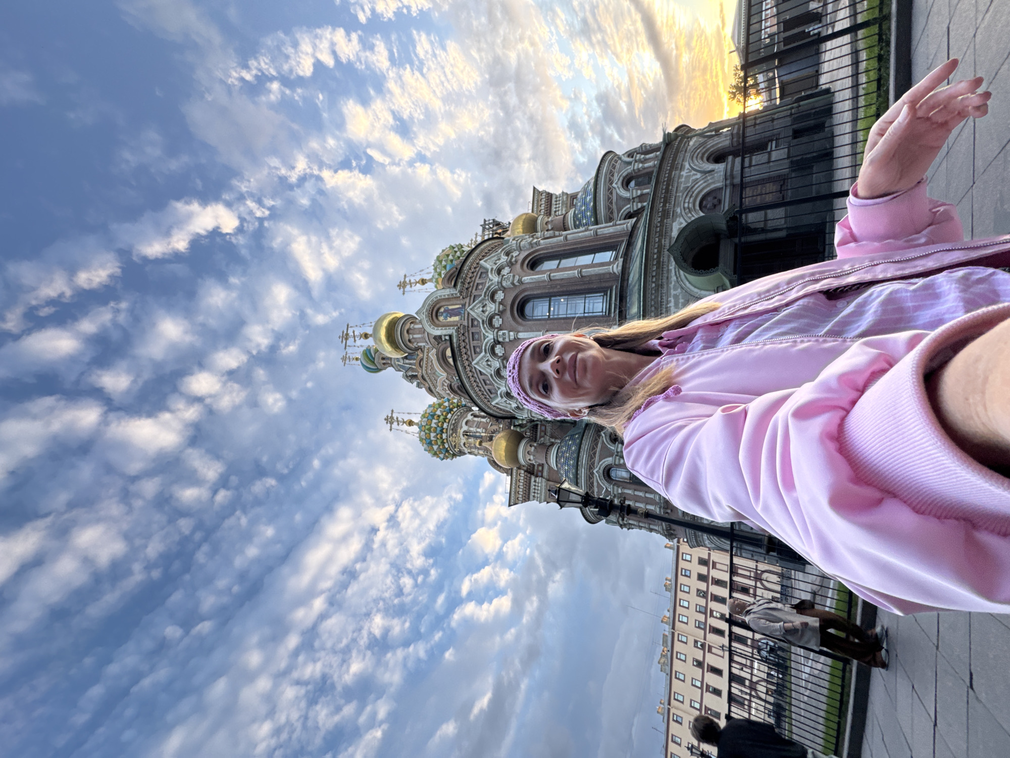The image shows a woman in a pink jacket and headband taking a selfie in front of the Church of the Savior on Spilled Blood in Saint Petersburg, Russia. The church is a prominent landmark with multiple golden and colorful domes. The sky above is blue with scattered white clouds, and the sun is setting, casting a warm glow on the scene. In the background, other people can be seen walking on a sidewalk, and a building with many windows is visible. The overall impression is a travel snapshot capturing a tourist moment at a significant architectural site.