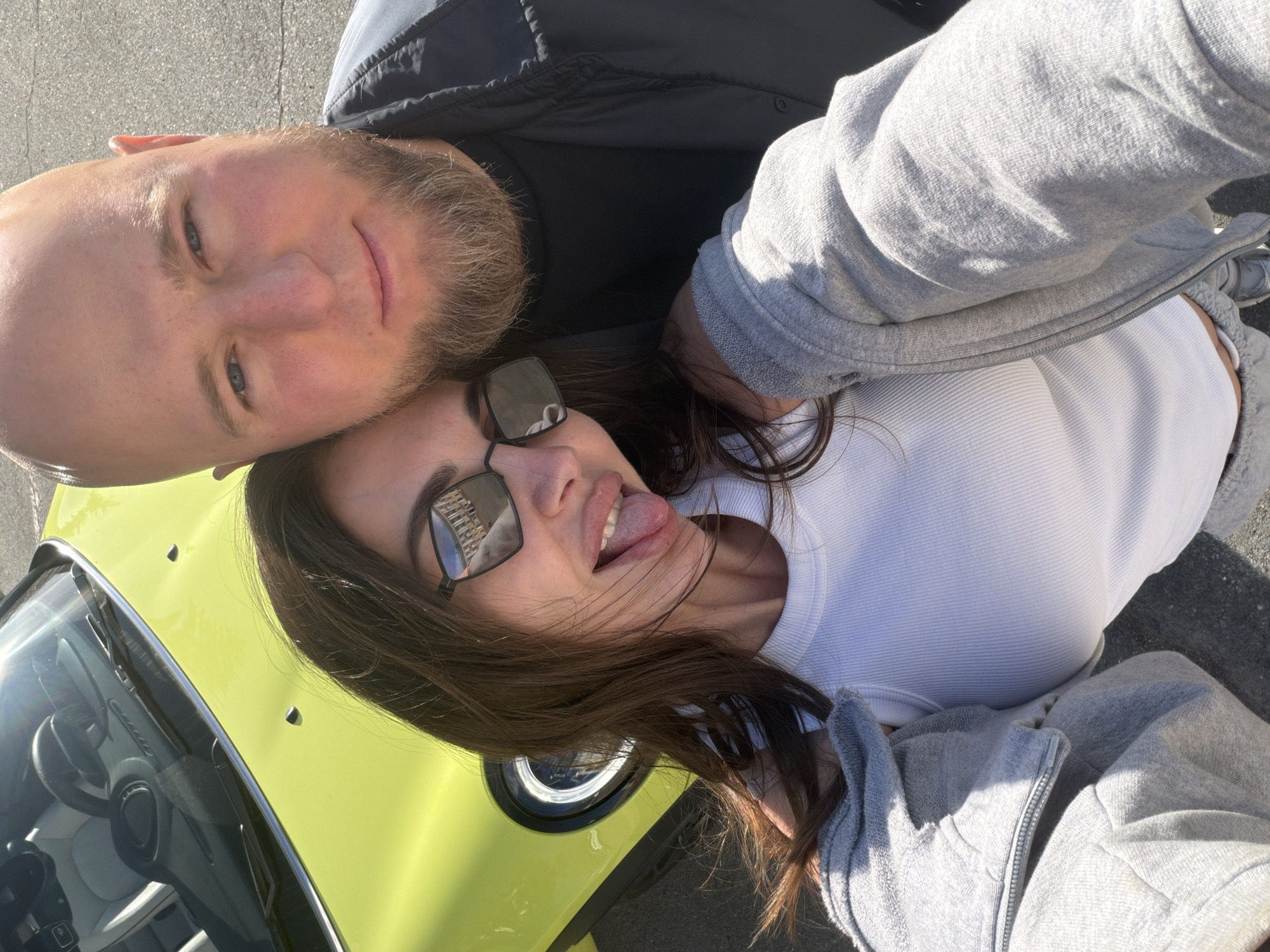 Two individuals are positioned close together, likely for a selfie. A bald man with a beard is near a woman with sunglasses who is sticking out her tongue. The front of a chartreuse-colored vehicle is visible, possibly part of a Mini Cooper, and the image appears to be taken outdoors. Without additional information or context, I cannot confidently assert it to be in Zheleznogorsk, Russia.
