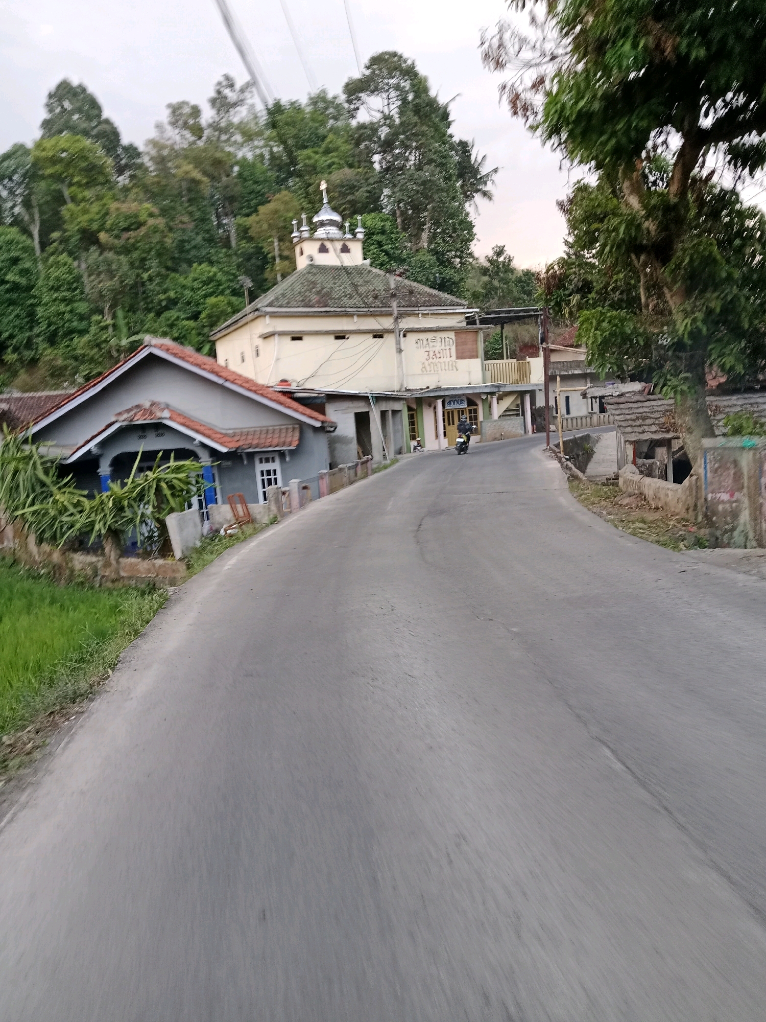 This image depicts a road in the city of Sirnagalih, Indonesia. The road is paved and curves to the right. On the left side of the road, there is a grey house with a red tile roof and a small lawn. Further down the road, there is a light yellow building with a domed roof and minarets, identified as "MASJID JAMI ABRUK" (Jami Abrue Mosque). A person is riding a motorcycle on the road in front of the mosque. The background is filled with lush green trees, suggesting a hilly or mountainous terrain. The sky is overcast.