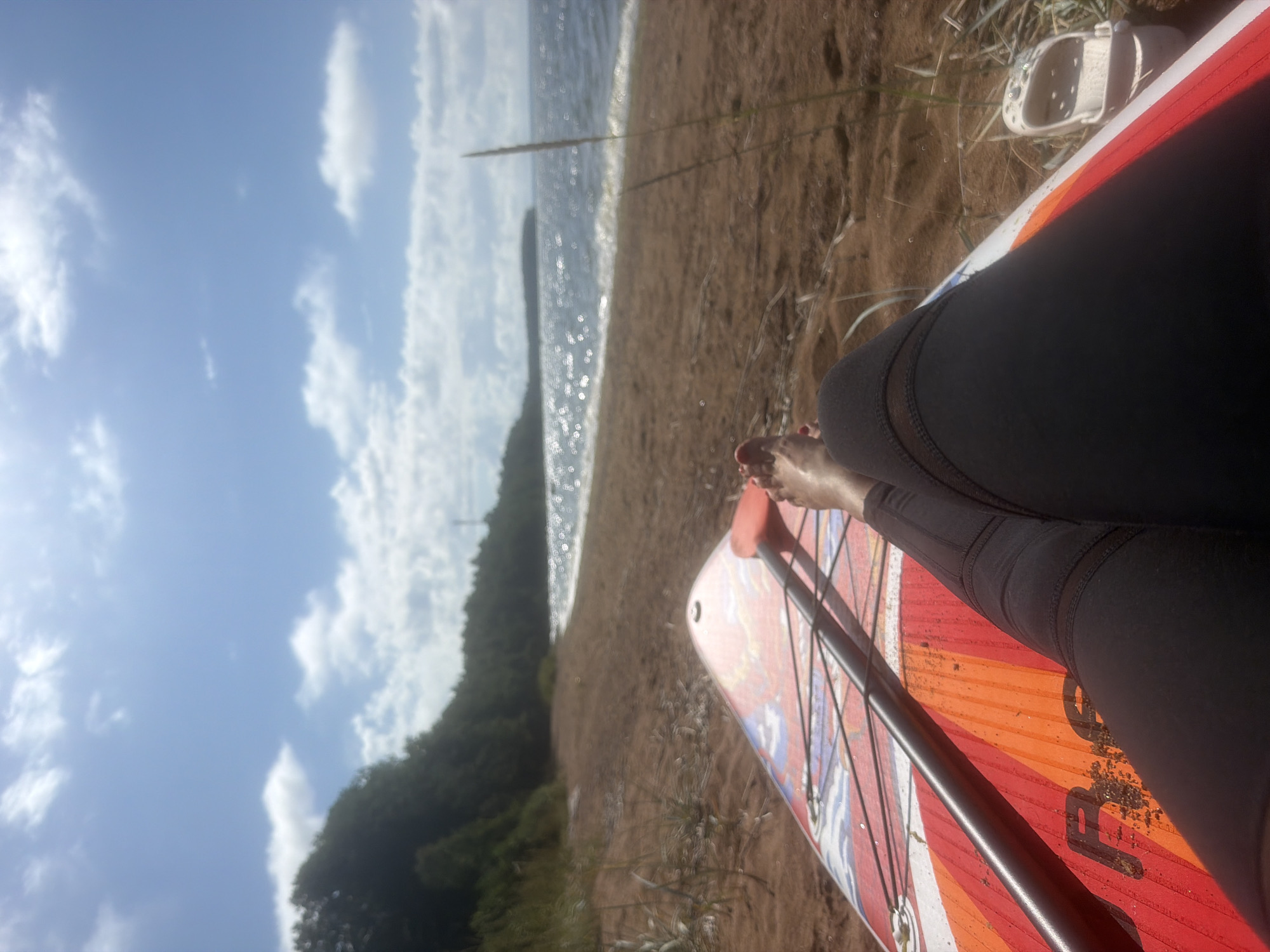 The image depicts a close-up, low-angle view of a person's legs and feet resting on a colorful paddleboard. The paddleboard is positioned on a sandy beach, with a paddle lying across it. Beyond the beach, a body of water, likely a lake or sea, is visible, with gentle waves lapping at the shore. The water sparkles under a bright, partially cloudy sky. A dense line of trees borders the beach in the distance, and a single tall stalk of grass grows in the foreground. A white Croc shoe is visible to the right of the paddleboard. The scene suggests a relaxed moment on a sunny day by the water. There are no specific indicators in the image that can confirm the location as Vyazy, Russia.