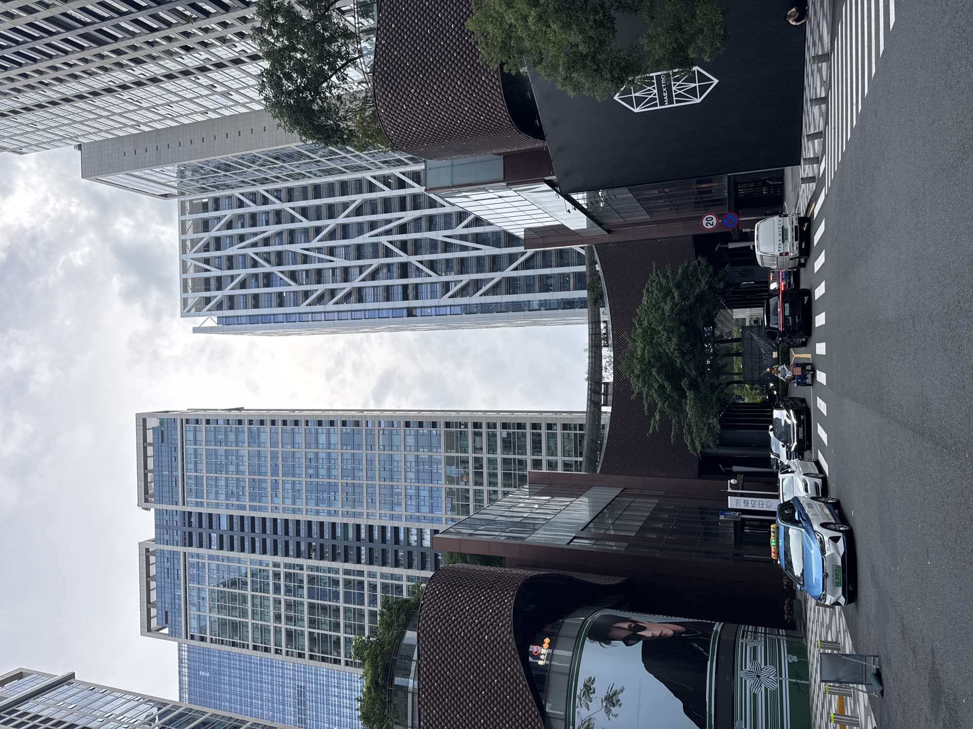This image shows a street scene in Futian District, Shenzhen, China, characterized by modern architecture and urban elements. Several tall buildings with glass facades and distinct architectural designs dominate the skyline. One building features a white exterior with a cross-lattice framework. A building with a dark, curved facade is also visible.
On the street level, cars are parked or driving. A blue and white taxi is at the forefront. Street markings, including a crosswalk and speed limit sign, are present. A pedestrian stands near the edge of the sidewalk. The environment suggests a bustling urban area with commercial and residential buildings.