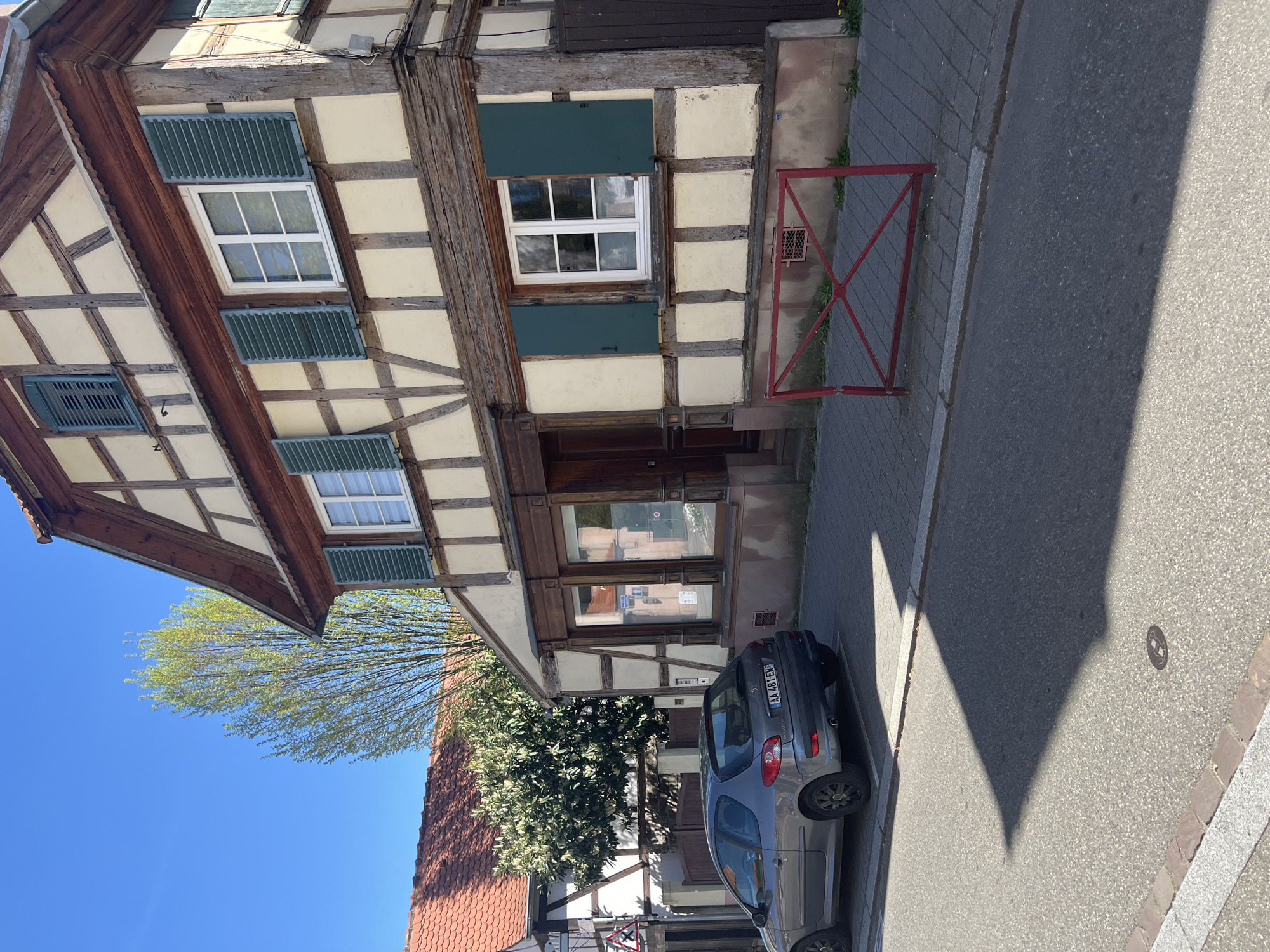 The image captures a scene in Schiltigheim, France. It shows a two-story timber-framed building with green shutters. A gray car is parked next to the building on the street. A red metal barrier is placed along the sidewalk in front of the building. The sky is clear and blue. A tree is on the left with light green foliage. A road with the shadow of a building can be seen. A section of a red-tiled roof is visible. The car has a license plate that appears to read "AA-481-EN."