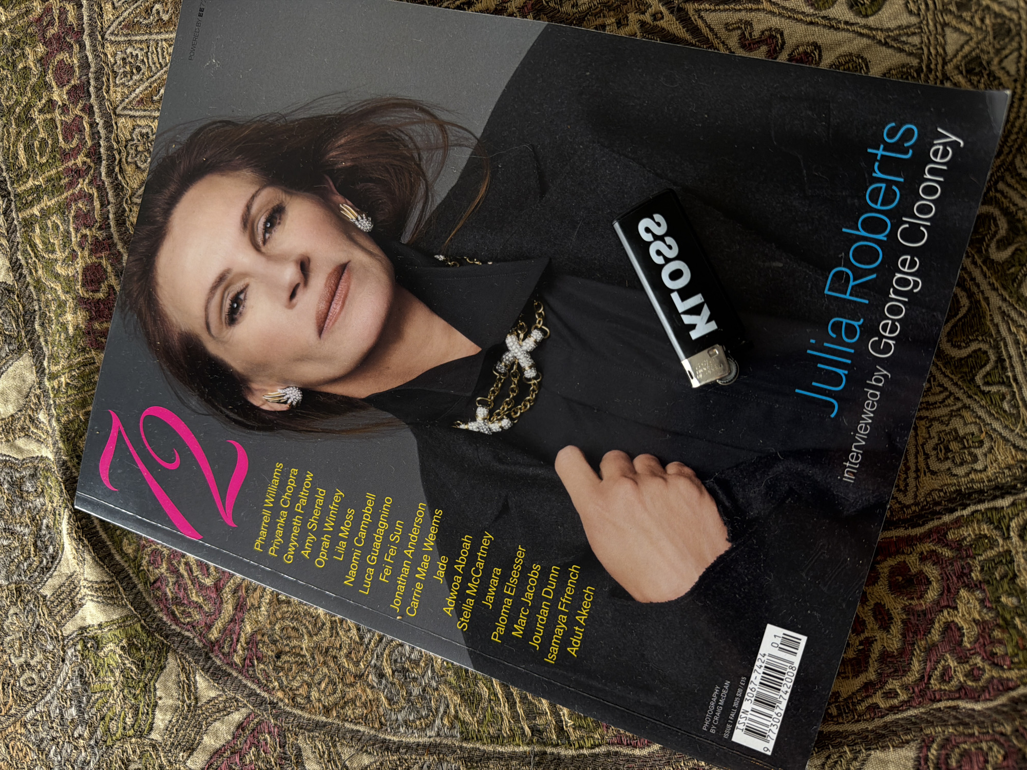 The image shows a close-up, overhead view of a magazine with Julia Roberts on the cover, lying on a patterned fabric surface. The magazine's title, "12", is prominently displayed in hot pink, and a list of names appears below it, including Pharrell Williams, Priyanka Chopra, Gwyneth Paltrow, Amy Sherald, Oprah Winfrey, Lila Moss, Naomi Campbell, Luca Guadagnino, Fei Fei Sun, Jonathan Anderson, Carrie Mae Weems, Jade, Adwoa Aboah, Stella McCartney, Jawara, Paloma Elsesser, Marc Jacobs, Jourdan Dunn, Isamaya Ffrench, and Adut Akech. Julia Roberts' face is visible, looking towards the viewer. A black lighter with the word "KR022" in white text is placed on the magazine cover, obscuring part of Julia Roberts' clothing. The bottom of the magazine shows publication details, including "PHOTOGRAPHY BY CRAIG MCDEAN" and "ISSUE 1 FALL 2025 $20 / £15". The fabric surface is a tapestry with a complex, ornate pattern in shades of brown, green, gold, and red. There is no indication of a specific location in London, United Kingdom.