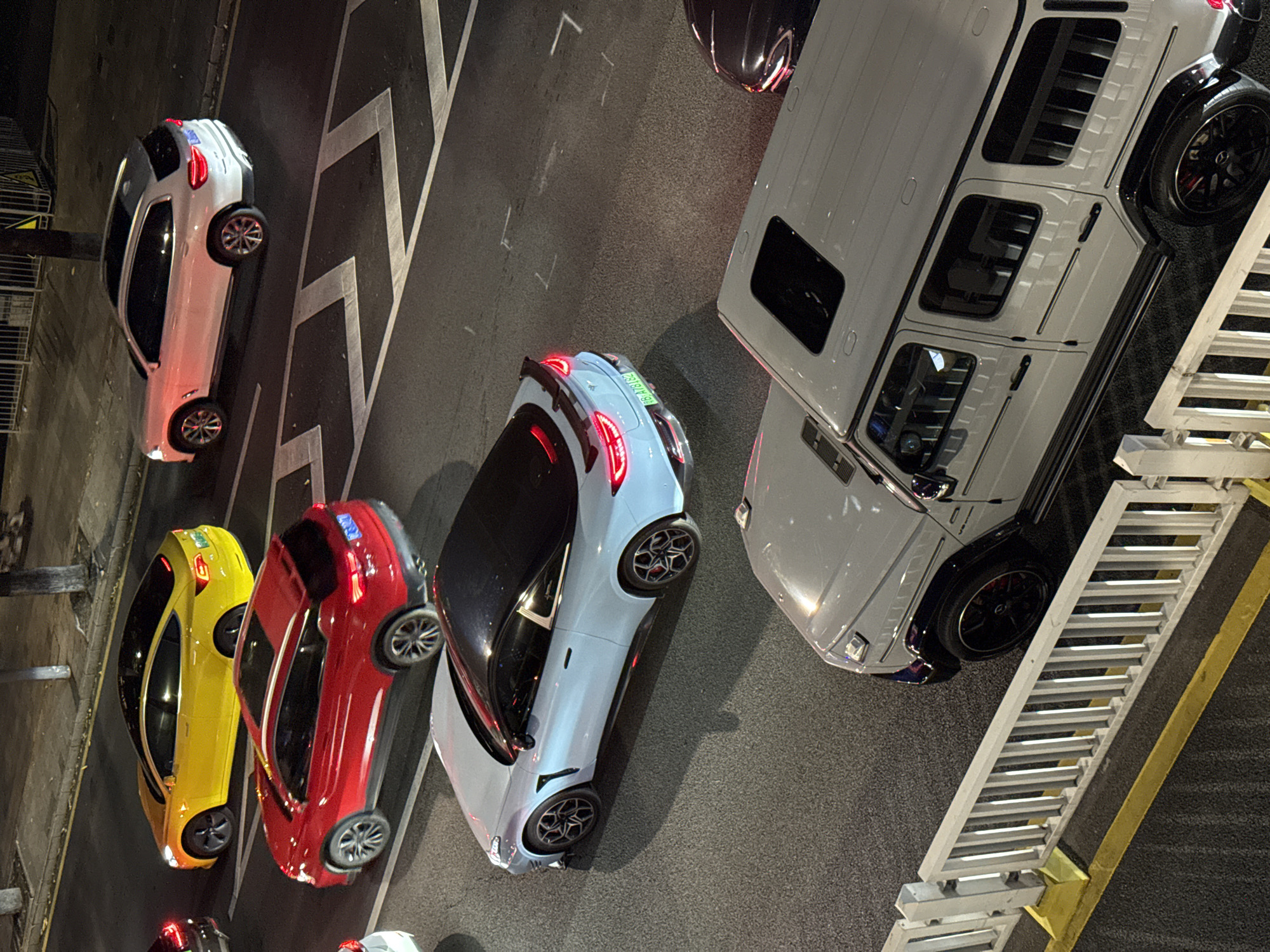 The image shows an overhead view of several cars parked on asphalt in what is potentially the Futian District in China. A white SUV, possibly a Mercedes-Benz G-Class, occupies a significant portion of the right side of the frame, positioned near a metal structure with yellow accents. To the left, there are a number of passenger vehicles in various colors, including yellow, red, and light blue. These are oriented perpendicularly to the SUV. Further to the left, there are other parked vehicles and structures. The scene is lit by artificial lighting, suggesting it is nighttime. Road markings are visible near the vehicles.