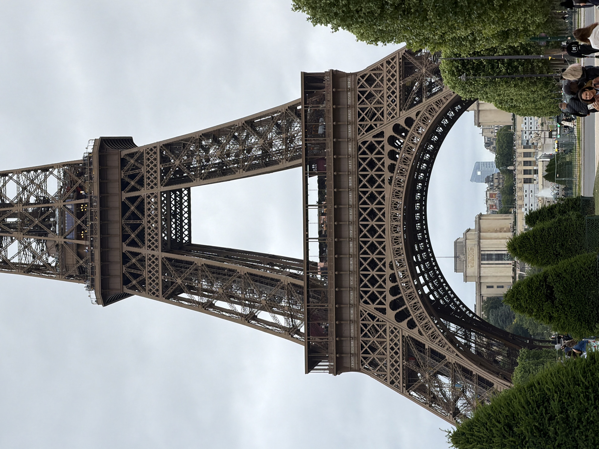 The image displays a prominent view of the Eiffel Tower in Paris, France, captured on an overcast day. The iconic iron lattice structure, with its dark metallic hue, dominates the frame, showcasing the intricate engineering and the grand arch of its base. The sky above is a uniform light grey, indicating cloudy weather.
In the foreground and mid-ground, lush green foliage from manicured bushes and trees lines pathways, suggesting the tower is situated within a park or garden area. Through the large arch at the base of the Eiffel Tower, a panoramic view of the Parisian cityscape is visible in the distance, featuring classic buildings and possibly the Palais de Chaillot.
Several people are scattered throughout the scene, primarily on the right side of the image. They appear to be visitors or tourists, some walking along the paths and others standing, likely admiring the landmark. A few individuals can also be faintly discerned on the tower's structure itself, possibly maintenance workers or visitors on the upper levels.
A notable detail of the Eiffel Tower's design is the frieze running along the first platform, where the names of eminent French scientists and engineers are engraved. Visible names include CAUCHY, BELGRAND, REGNAULT, FRESNEL, DE PRONY, VICAT, EBELMEN, COULOMB, POINSOT, FOUCAULT, DELAUNAY, MORIN, HAUY, COMBES, THENARD, ARAGO, POISSON, and MONGE. The scene depicts typical daytime tourist activity around one of the world's most famous monuments.