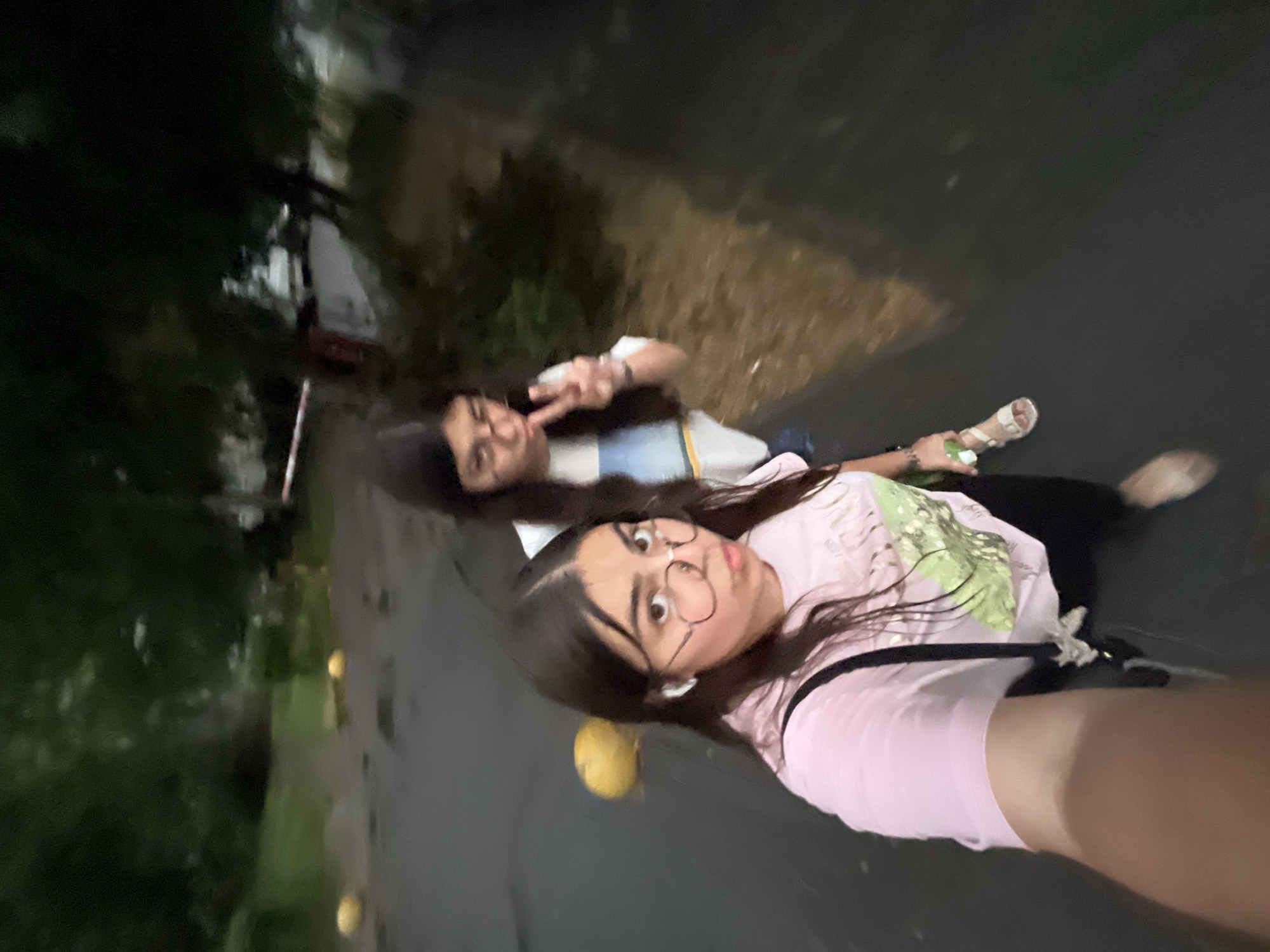 Two young women are walking on a wet asphalt surface, likely a street or sidewalk, on a cloudy day. The woman in the foreground is wearing a pink t-shirt and black pants, with round glasses and white wireless earbuds. The woman behind her is wearing a white t-shirt with a striped design and is holding up two fingers in a peace sign. Yellow bollards are visible in the background. Trees and foliage line the area, suggesting a park or a street with greenery. The lighting indicates it may be overcast or dusk. The presence of wet asphalt suggests recent rain. The image is taken from a selfie perspective, with the camera held by the woman in the foreground.