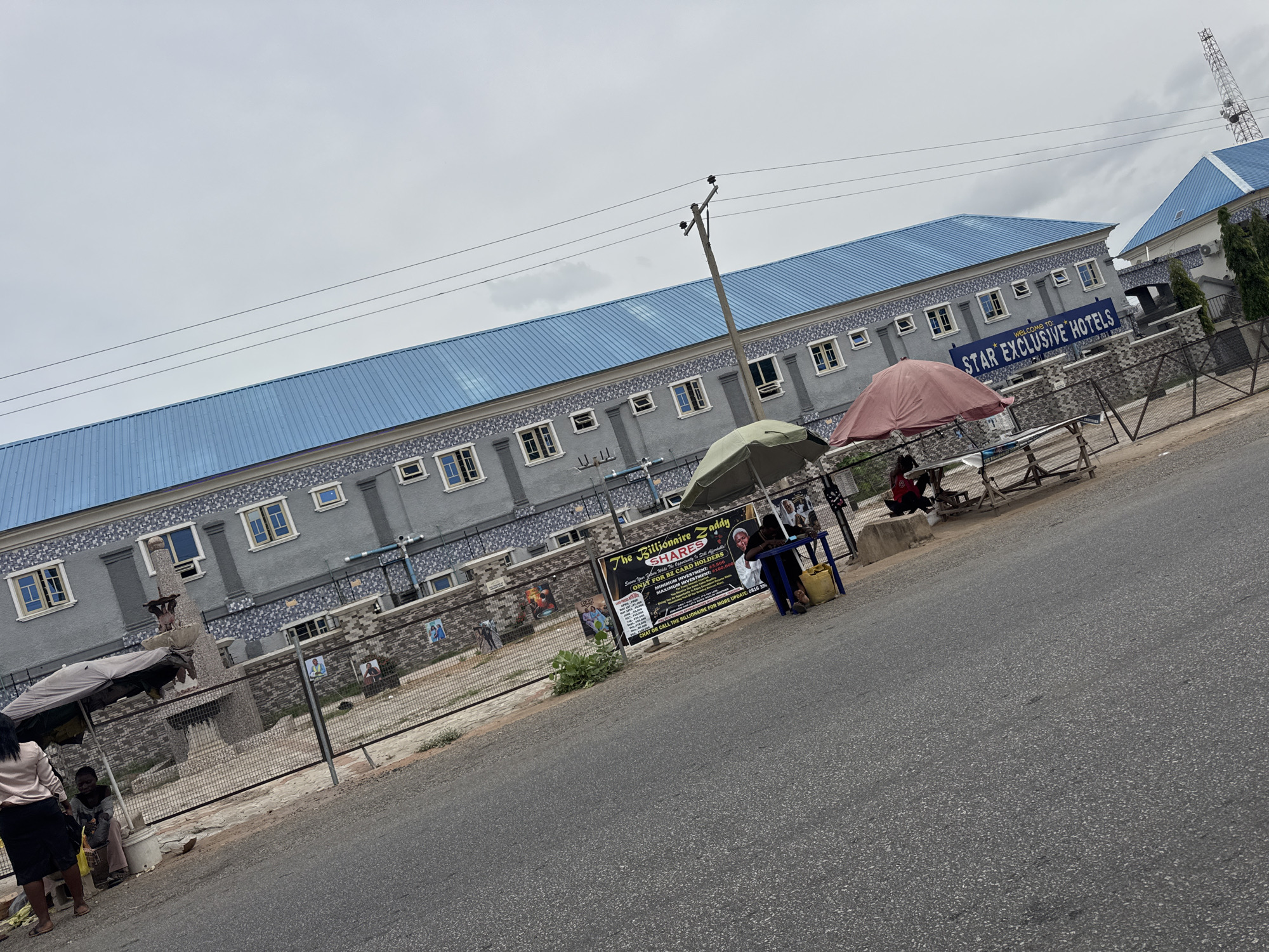 This media file captures a street view of "STAR EXCLUSIVE HOTELS" in Jalingo, Nigeria. The hotel building features a distinctive light blue, corrugated metal roof and a grey facade decorated with a repeating geometric pattern around the windows. Several windows are visible, some open and some closed, suggesting the hotel is operational. In front of the hotel, a metal fence encloses the property.
Outside the fence, several people are present. Two individuals are seated under a light-colored umbrella near a table, appearing to be engaged in an activity like selling or discussing something. Another person, a woman, is standing on the left side of the frame, facing away from the camera, dressed in a light-colored blouse and dark skirt. A young person in a brown shirt is sitting near a bucket under a darker umbrella. A wooden table with a red umbrella is set up further down, with another person seated there. Various signs are visible, including one that reads "The Billionaire addy SHARES" and "WELCOME TO: STAR EXCLUSIVE HOTELS". The sky is overcast and appears to be daytime, possibly late afternoon or early morning, given the diffused light. The overall scene suggests a bustling street in a Nigerian city, with commercial activity taking place outside the hotel.