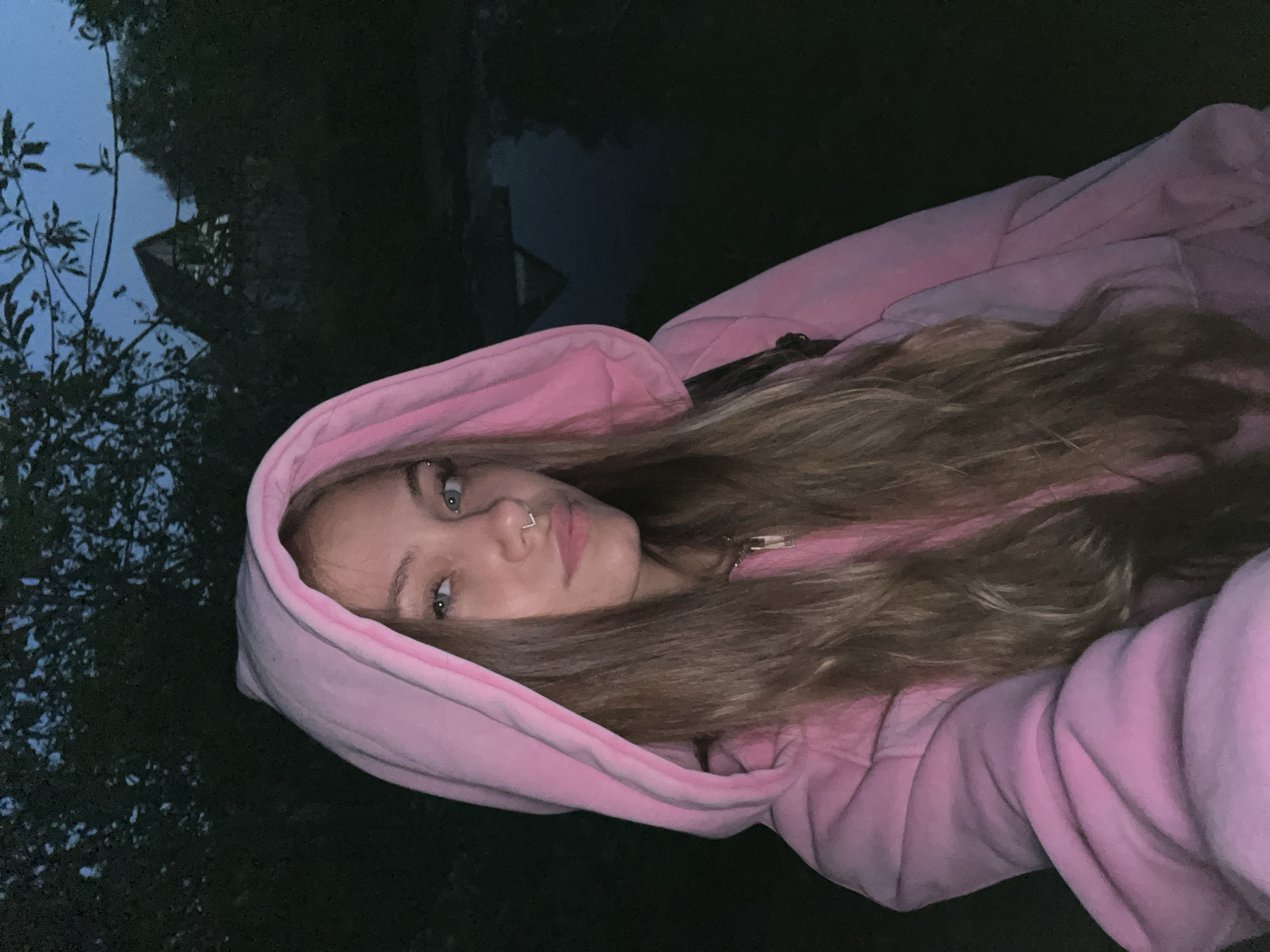 A young woman with light skin, long brown hair, and a nose piercing is centered in the image, wearing a pink hooded sweatshirt. She is looking directly at the camera with a neutral expression. The background is dark, but appears to be an outdoor environment with trees and what may be water. The location appears to be Фоки, Russia, based on the surrounding environment and context clues. The time of day appears to be evening or night due to the low lighting conditions.
