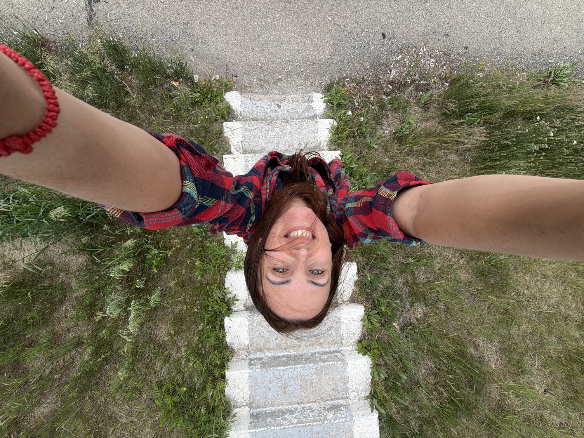This is a high-angle, selfie-style photo taken outdoors, likely in a rural or natural setting, possibly near Nikolaevka, Russia. The subject is a woman with long, dark brown hair and blue eyes, who is smiling broadly at the camera. She is wearing a red, blue, and black plaid shirt and a red beaded bracelet on her left wrist.
The woman is positioned on a set of old, weathered concrete steps, surrounded by tall, dry grass and some scattered weeds. Above the steps, there appears to be a paved area, possibly a road or path. The overall scene suggests an overcast or moderately lit day, with no strong shadows indicating a specific time of day. The dry grass hints at either a dry season or a less fertile area.
The perspective of the photo, looking straight down at the woman as if from above, creates a unique and somewhat disorienting viewpoint. Her expression is cheerful and energetic, suggesting she is enjoying herself or the experience of taking the photo. There is no visible text in the image.