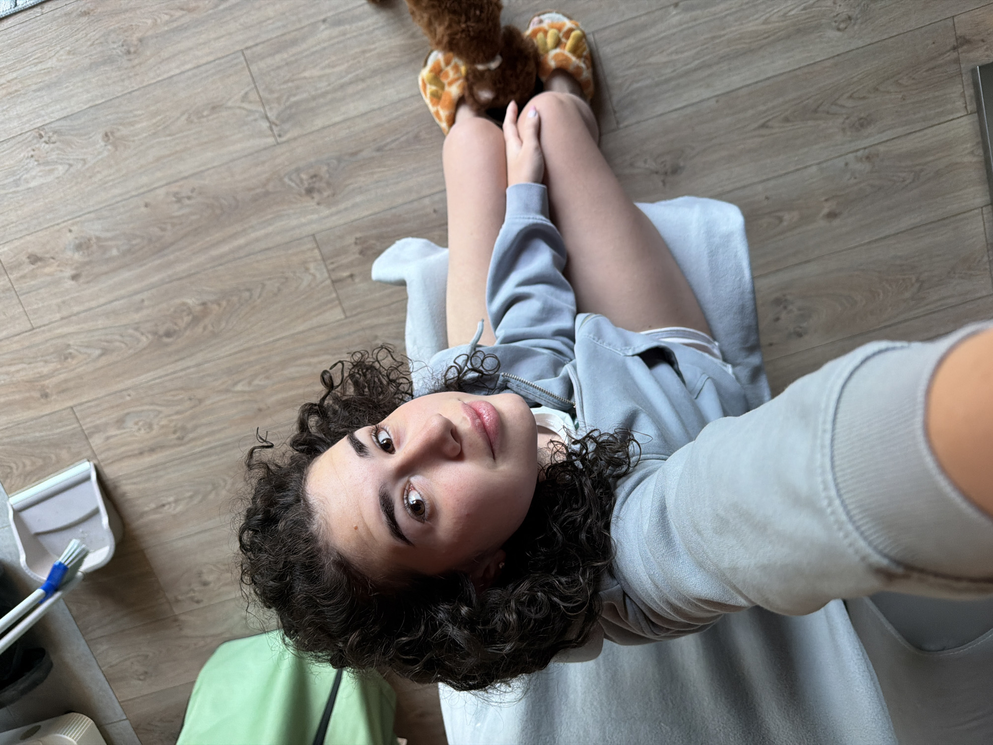 A young Caucasian woman with dark curly hair, wearing a light blue hoodie and slippers, is seated on a light-colored piece of furniture. She is looking up at the camera, holding it to take a selfie. Her legs are crossed, and she is wearing light gray socks. The background consists of a light brown wooden floor and a green cloth bag. A dustpan and brush are visible in the upper left corner of the image. The scene appears to be indoors, possibly in a residential setting. The image does not provide any information about the specific location in Sofia, Bulgaria.