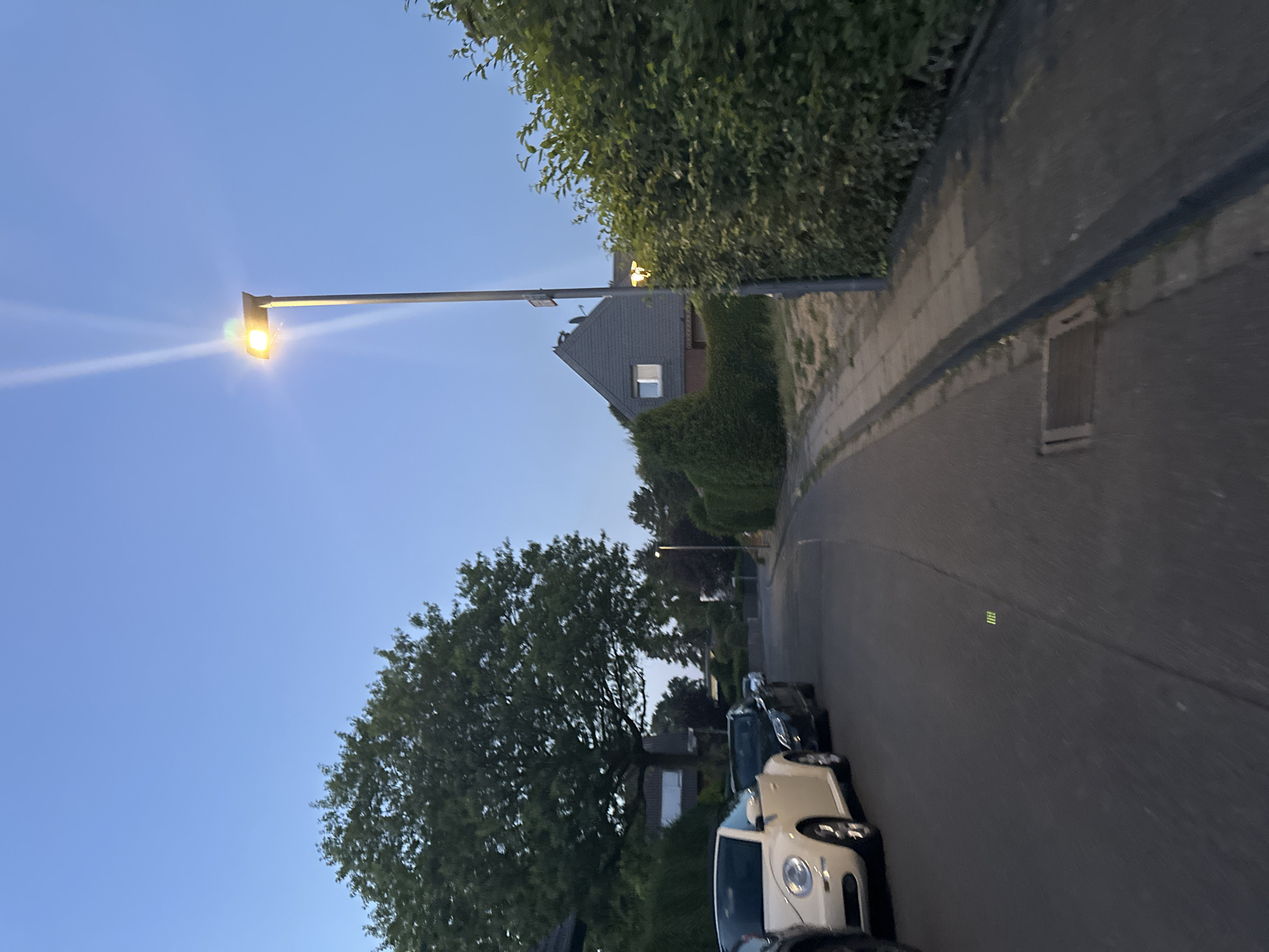 The image depicts a street scene in Krefeld, Germany, during twilight. A street lamp on the right side of the frame illuminates the area. In the foreground, a paved road is visible, with a sidewalk bordering it. Several parked cars are on the left side of the road, including a white convertible and a dark sedan. Behind the cars, mature trees and hedges line the street. Residential buildings are visible in the background, with a gray-roofed house prominently featured. The sky is a clear, pale blue, indicating the early evening or late twilight hours.