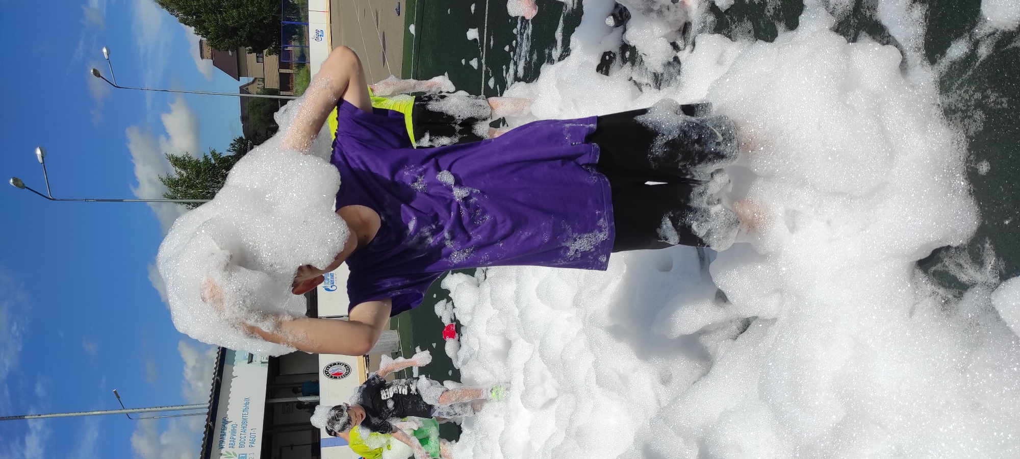 In Noviy, Russia, on a sunny day, a young boy in a purple t-shirt and black shorts is immersed in a foam party. He is holding a large mass of foam over his head with both hands, obscuring his face. The foam party is taking place outdoors on what appears to be a sports field. Other children are visible in the background, also playing in the foam. One boy in a black t-shirt and shorts is running through the foam, while another child in a yellow t-shirt is partially visible. The environment includes a building with signage in Russian, streetlights, trees, and a blue sky with white clouds. The scene suggests a recreational event for children.
