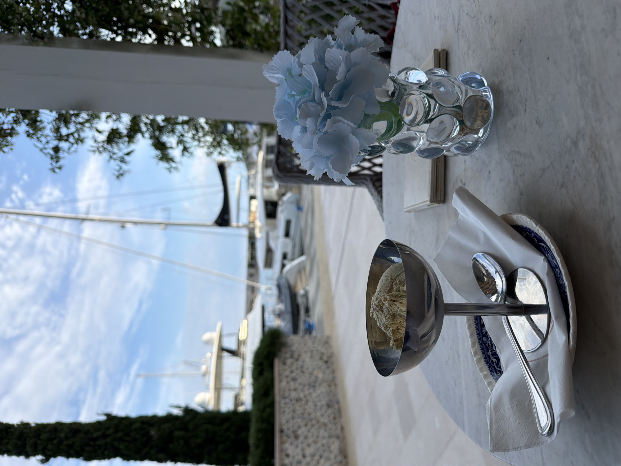 A high-angle, rotated, outdoor shot shows a dessert setting on a white marble table, possibly in Radovići, Montenegro, considering the marina-like setting and presence of boats. The primary subject is a silver bowl containing a beige-colored dessert, possibly ice cream or sorbet, on a silver pedestal stem sitting in a white cloth napkin on a blue-and-white patterned plate. A fork and a spoon rest on the napkin next to the bowl. To the right of the dessert, there's a small, glass vase filled with light blue flowers, and behind the vase, there are beige notepads. The background shows boats docked at a marina and greenery and a white structure under which the table is likely located. The sky is visible in the background, suggesting daylight. The scene gives an impression of a meal at an upscale outdoor dining location.