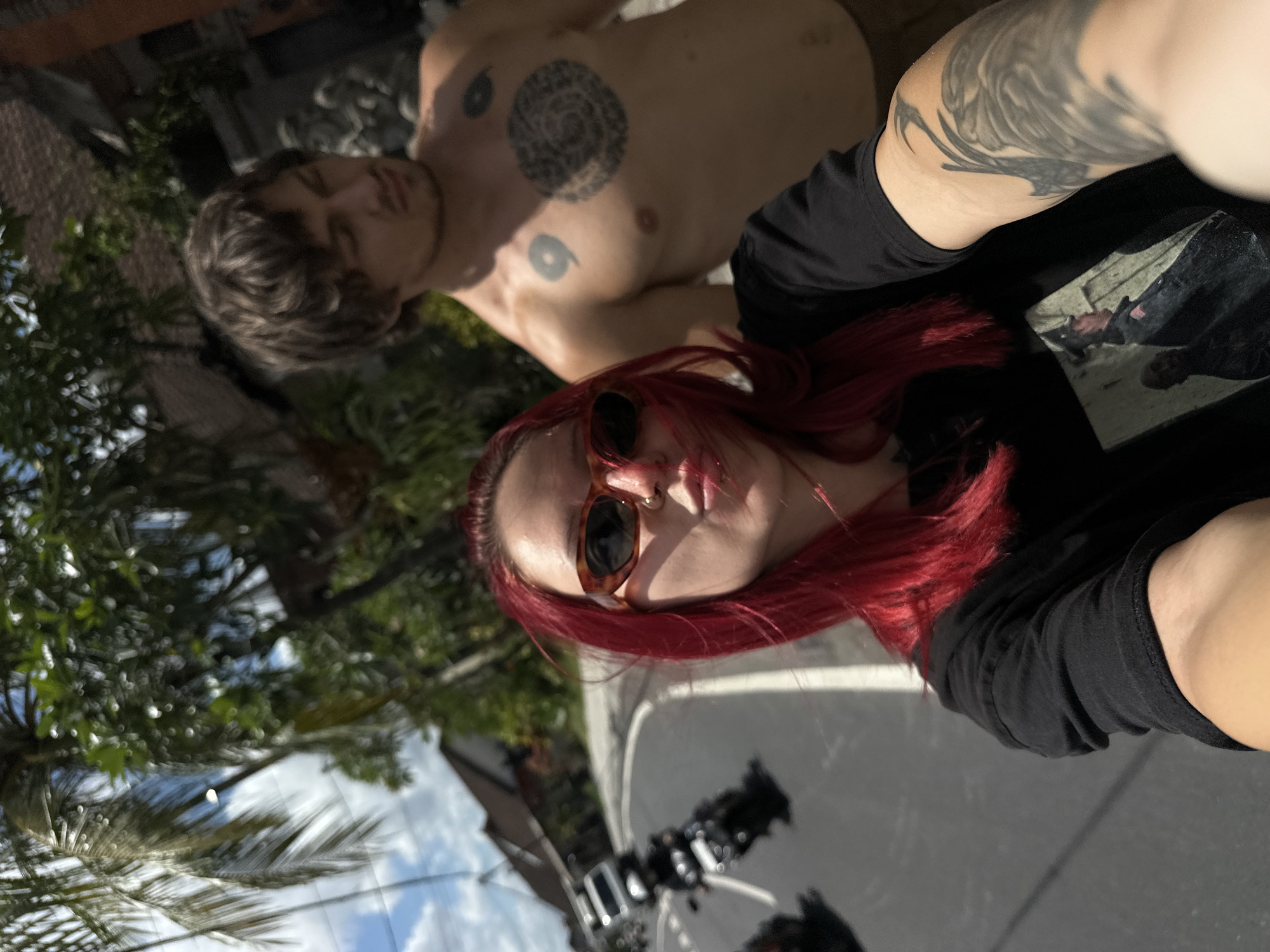 Two people are in the image. One shirtless person with tattoos is behind a person with red hair and sunglasses wearing a black shirt, who appears to be holding the camera. There are trees and buildings in the background. The location is likely North Kuta, Indonesia.
