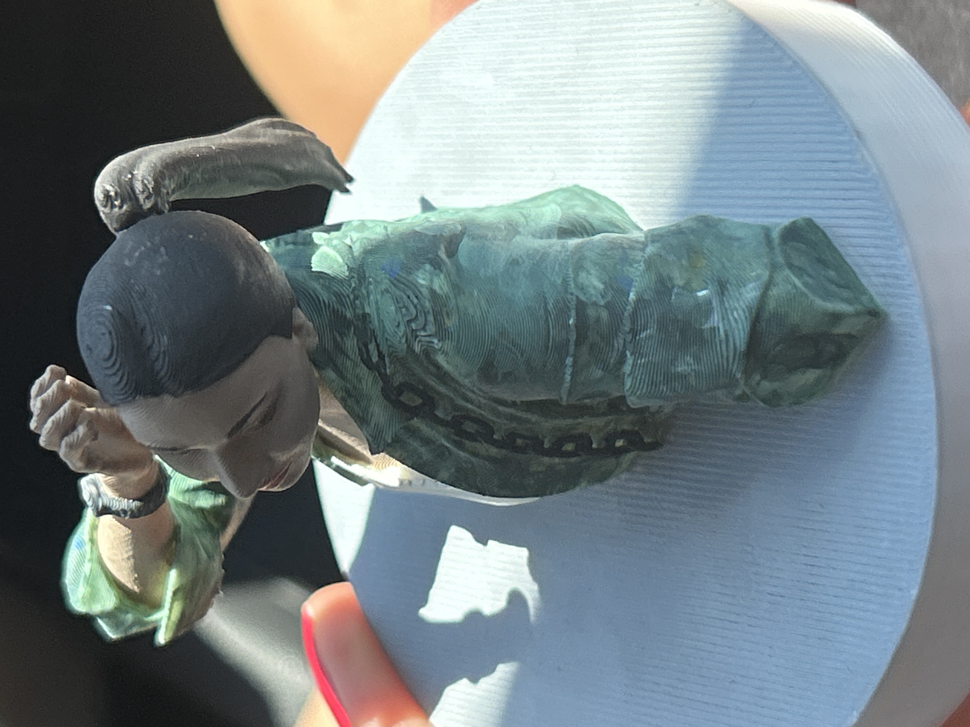 The image shows a close-up, top-down view of a 3D-printed miniature figure of a person, possibly a historical figure, placed on a light blue, circular base. The figure is depicted from the chest up, wearing a green jacket with intricate detailing and a white undershirt. The figure has dark hair styled in two pigtails and is looking to the left with its right arm raised. The base has a ribbed texture. A person's hand with red-painted fingernails is visible in the lower left corner, casting a shadow on the base. The background is dark and out of focus. There is no information about the location of this image in Lozen, Bulgaria.