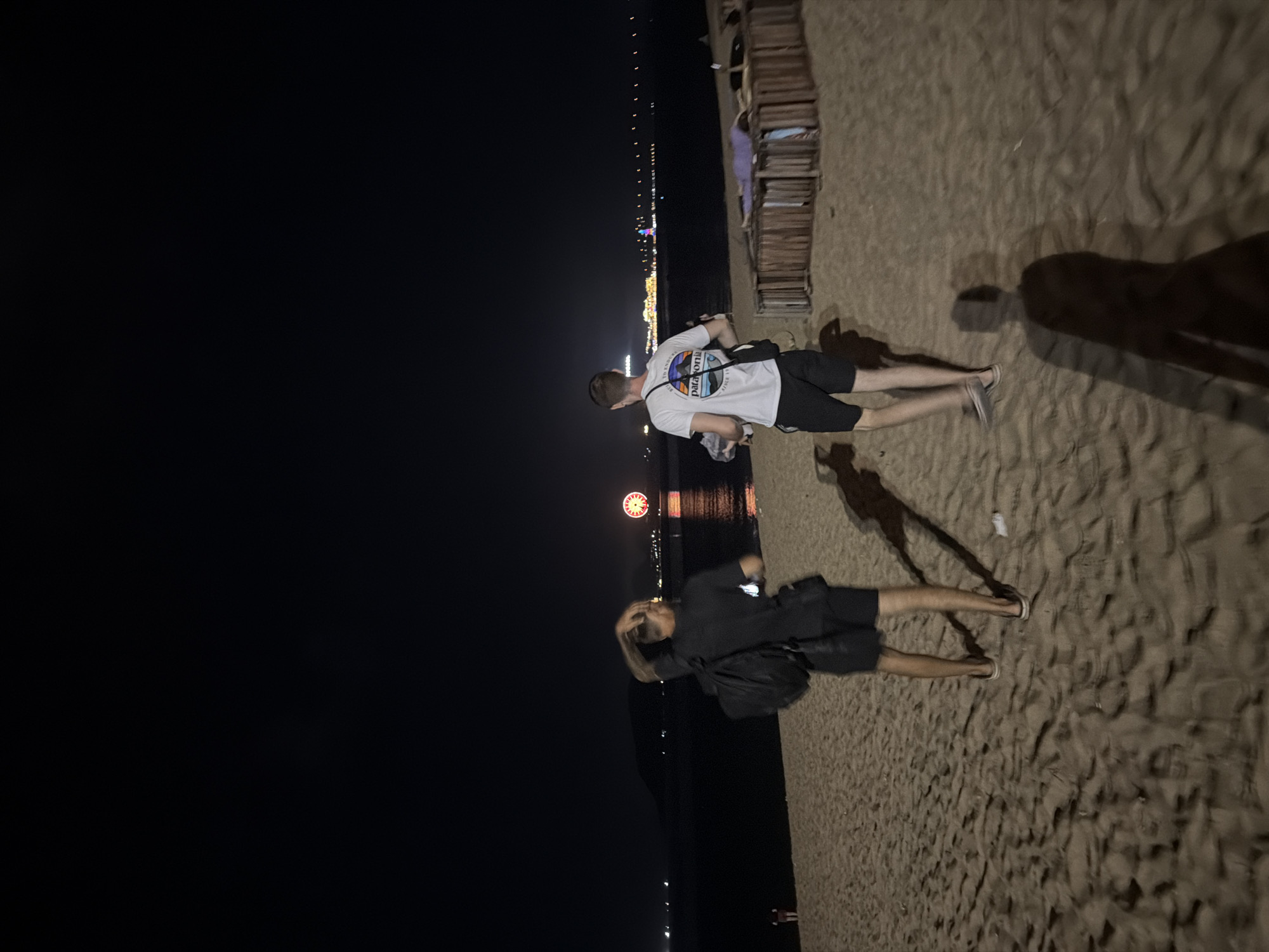 Two men are walking on a sandy beach at night in Nha Trang Ward, Vietnam. The man on the left is wearing a black t-shirt and black shorts and has a backpack. The man on the right is wearing a white t-shirt with a graphic print and black shorts. In the background, there is a body of water with a Ferris wheel and lights from the city visible on the horizon. The beach is illuminated by artificial lights and the glow from the city.