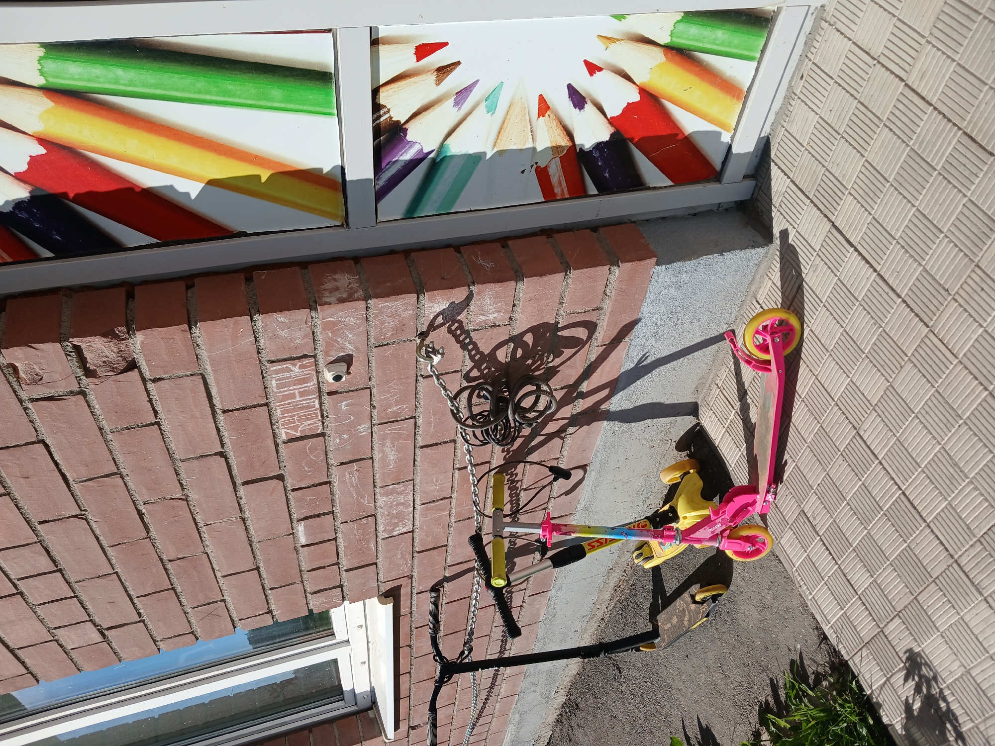 The image shows two scooters parked outside a brick building, possibly in a city like Yekaterinburg, Russia. A black scooter with yellow handles is on the left, and a pink and yellow three-wheeled scooter is on the right. Both scooters are chained to the wall. The building has a white-framed window and a display with colorful pencils. The ground is paved with light-colored tiles.