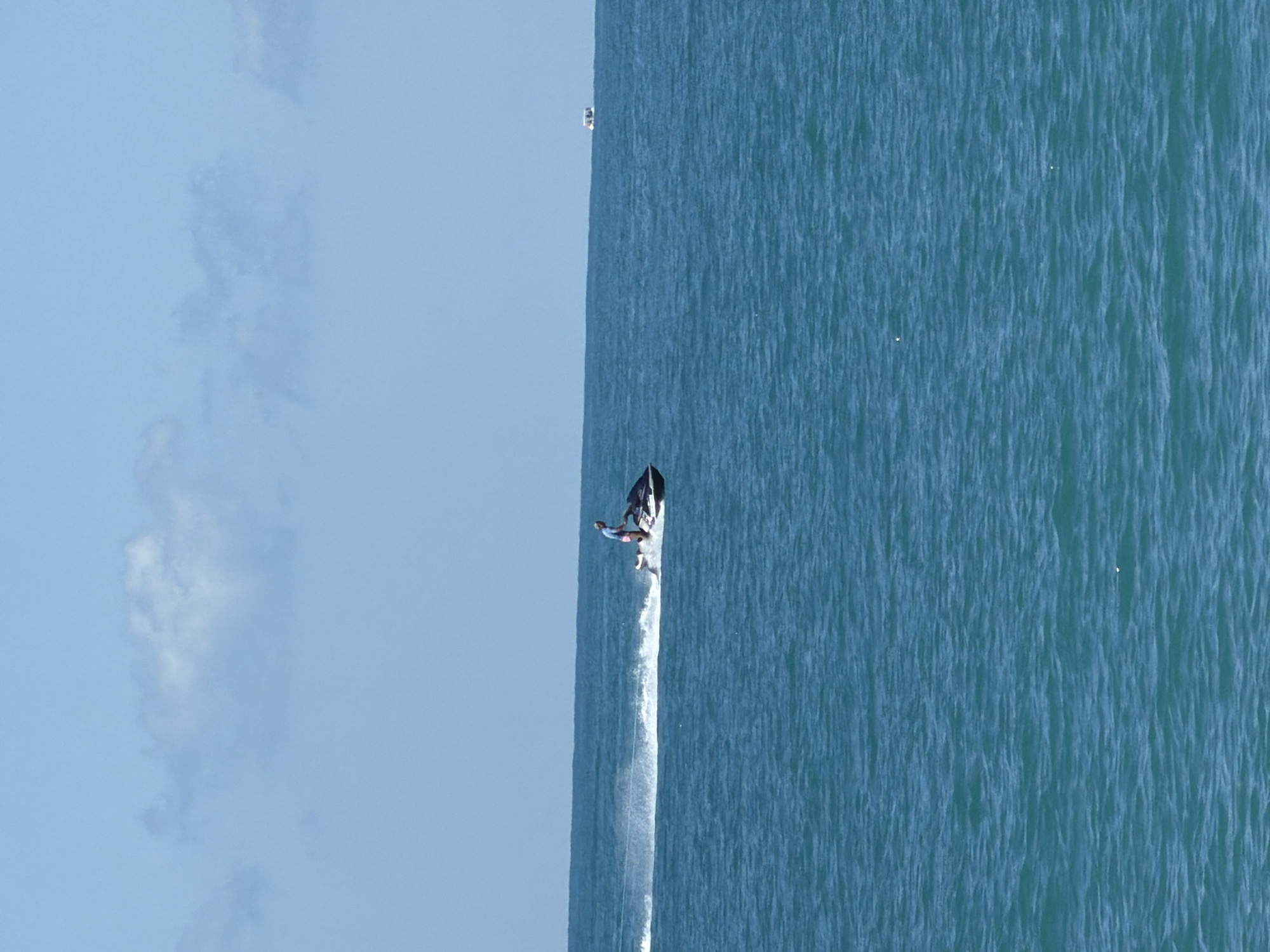 A person is riding a jet ski across the turquoise water of the sea. The jet ski is leaving a white wake behind it. The sky is blue with a few white clouds. In the distance, there are two small boats on the horizon. The scene appears to be a recreational area, possibly in Sochi, Russia, given the clear blue sky and vibrant sea colors often associated with the Black Sea coast.