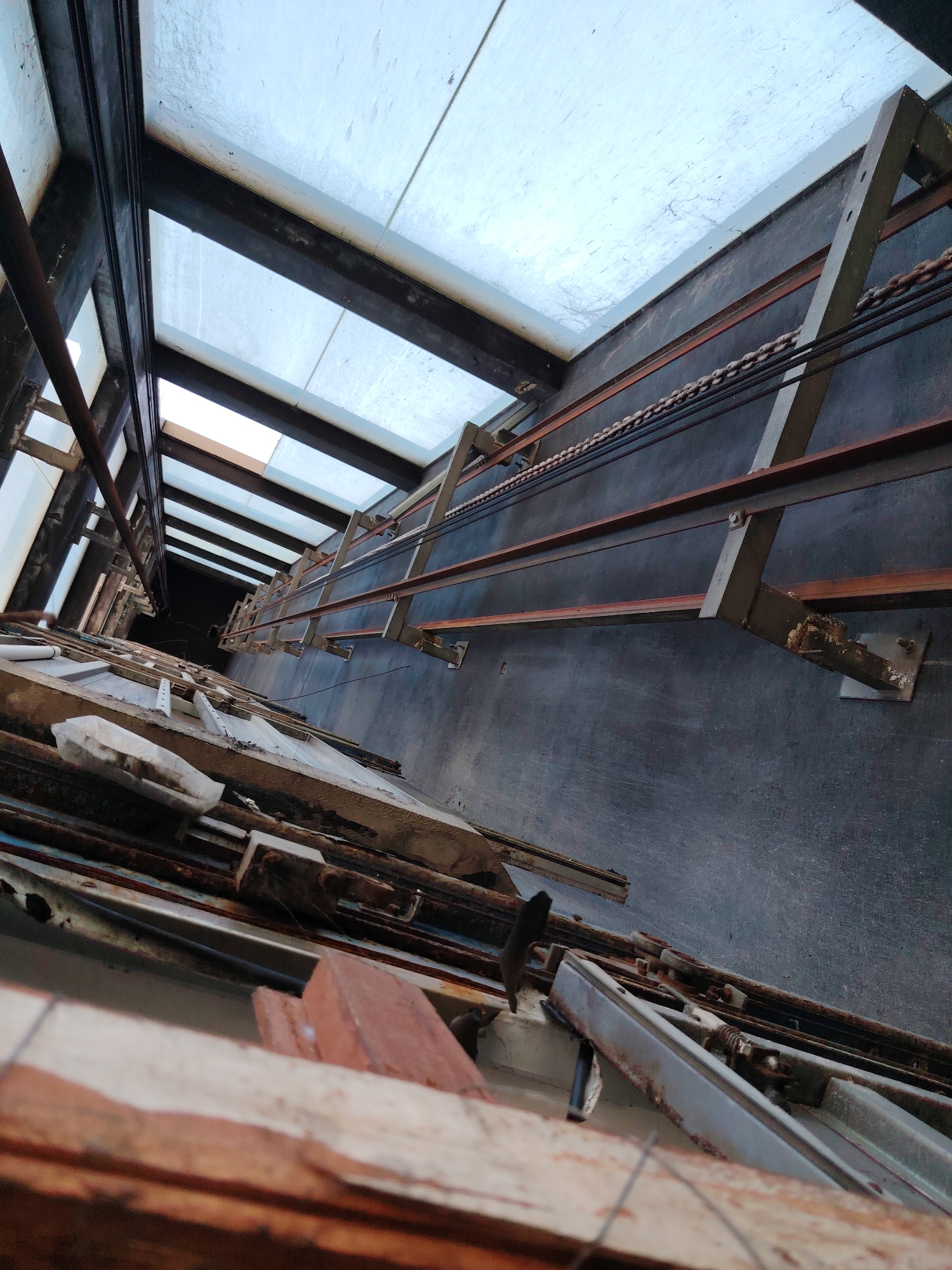 This image depicts the interior of an abandoned elevator shaft looking upwards. The shaft is dark and appears to be several stories deep, with rusted metal beams and cables running along the walls. The ceiling is made of translucent panels, allowing natural light to filter in. Debris is scattered throughout the shaft, including broken wooden planks and discarded metal parts. The perspective is from the bottom of the shaft, looking up towards the light. There are no people or animals visible in the image. The overall impression is one of decay and disuse.