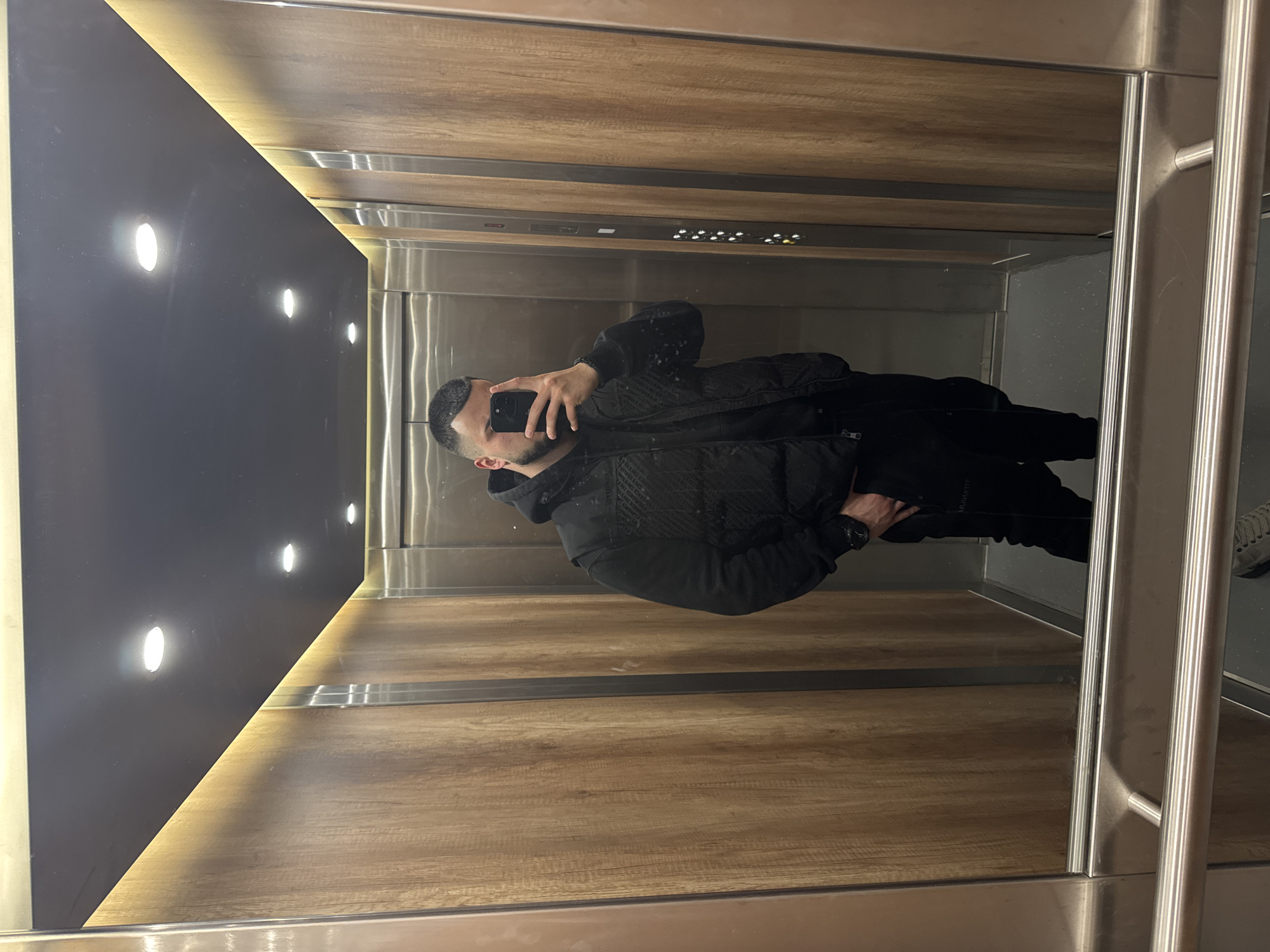 The image shows a man taking a selfie in an elevator. The elevator has wood paneling on the walls and a dark panel with lights on one side. The floor appears to be the same wood paneling as the walls. The elevator doors are made of stainless steel. The man is wearing a black jacket and black pants. The image does not provide enough information to identify the specific location within Sofia, Bulgaria.