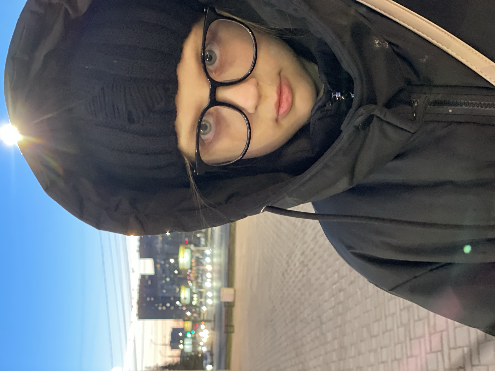 A young person, likely female, is featured in a close-up, upward-angled selfie taken outdoors in an urban setting during dusk or dawn. She wears black-rimmed glasses, a black knitted beanie, and a black hooded winter jacket, with the hood pulled up around her head. Her light-colored eyes look directly at the camera, and she has a neutral expression. A light-colored strap, possibly from a bag, crosses over her right shoulder.
The background on the left reveals a clear blue sky with a prominent, bright sunburst, indicating the sun is low in the sky. The right side of the background shows a city street with blurred buildings and numerous artificial lights from streetlamps and distant structures, suggesting the presence of traffic or active urban life in Izhevsk, Russia. The foreground consists of a paved sidewalk made of light-colored, rectangular blocks. The clothing suggests cold weather. There is no visible text in the image.