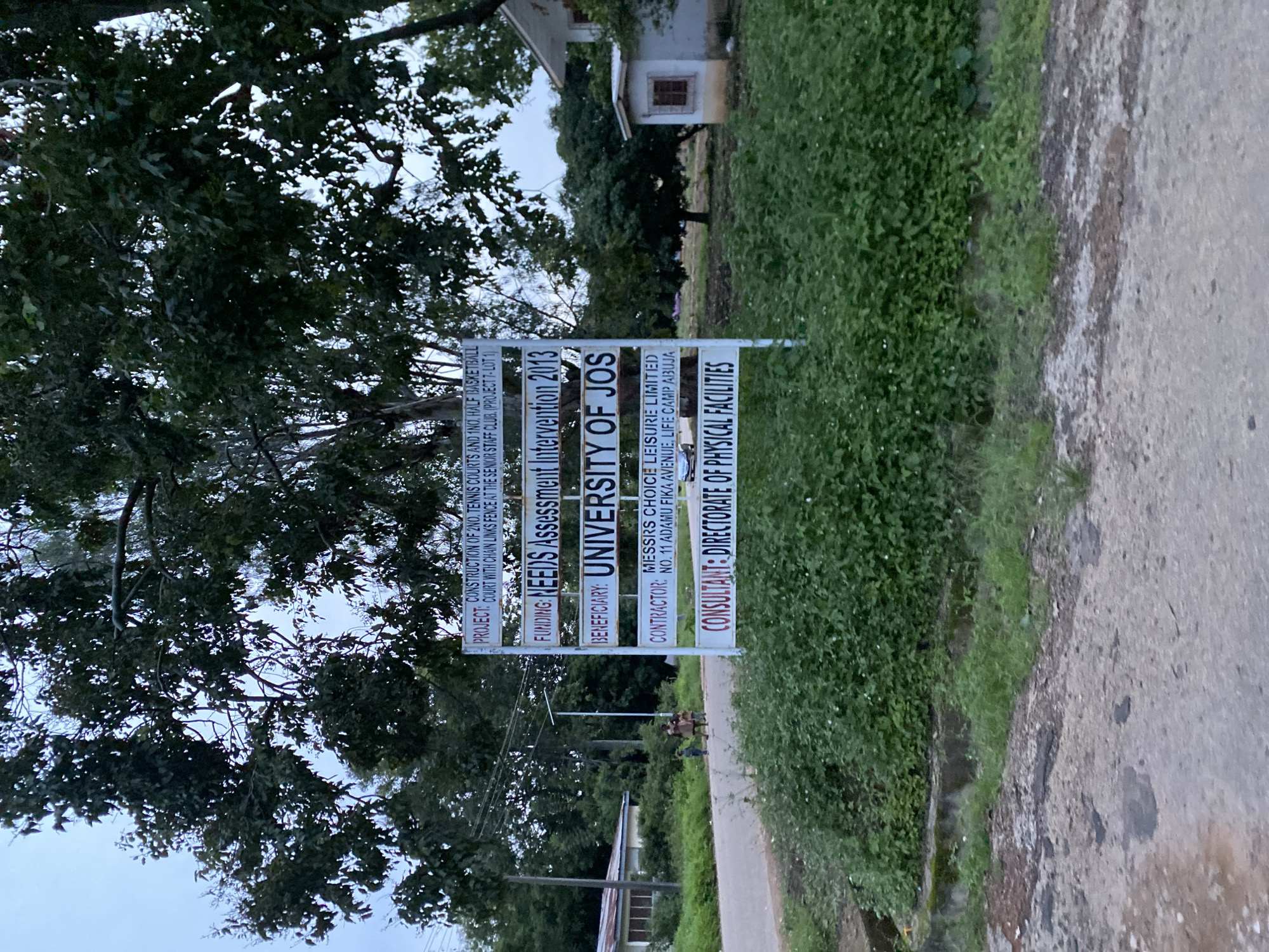 This image features a signpost with white panels and red lettering, providing information about a project. The sign is situated outdoors, near lush green vegetation and a paved path. Trees with dense foliage partially obscure the background, suggesting a natural or park-like setting. The sky appears overcast, indicating a cloudy day.
The text on the sign indicates that the project involves the "CONSTRUCTION OF 2NO. TENNIS COURTS AND 1NO. HALF BASKETBALL COURT WITH CHAIN LINKS FENCE AT THE SENIOR STAFF CLUB." The "FUNDING" is identified as "NEEDS Assessment Intervention 2013," and the "BENEFICIARY" is the "UNIVERSITY OF JOS." The "CONTRACTOR" is listed as "MESSRS CHOICE LEISURE LIMITED," with an address at "NO. 11 ADAMU FIKA AVENUE, LIFE CAMP ABUJA." The "CONSULTANT" for the project is the "DIRECTORATE OF PHYSICAL FACILITIES."
In the background, a few people are visible, standing on the path. They are not the primary focus of the image, which is dominated by the sign and the surrounding greenery. The overall impression is one of an official project being documented, likely at an educational institution in Jos, Nigeria, given the mention of the University of Jos. The lighting suggests it is daytime, possibly late afternoon or early morning, given the diffused light.