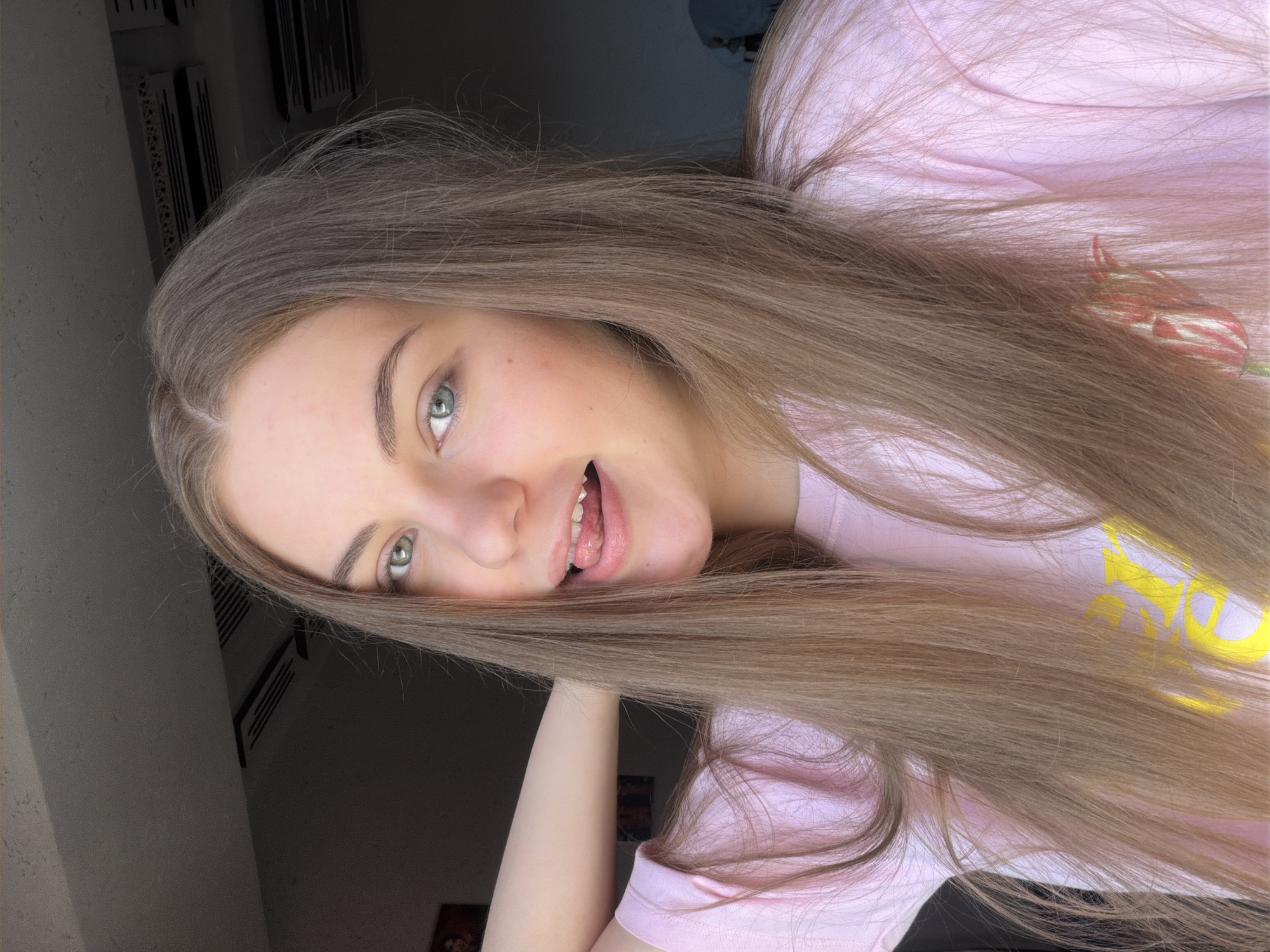 The image shows a medium shot of a young woman in a pink t-shirt, posing with her hand behind her neck. She has long, light brown hair that falls over her shoulders and chest. Her eyes are a striking blue-green, and her mouth is slightly open, with her tongue peeking out. The background is dimly lit, with what appears to be ceiling vents visible in the upper portion of the frame. A framed picture is partially visible on the wall to the left. The overall impression is that of a selfie taken indoors. There is no specific information in the image to indicate the location in the city of Dnipro, Ukraine.