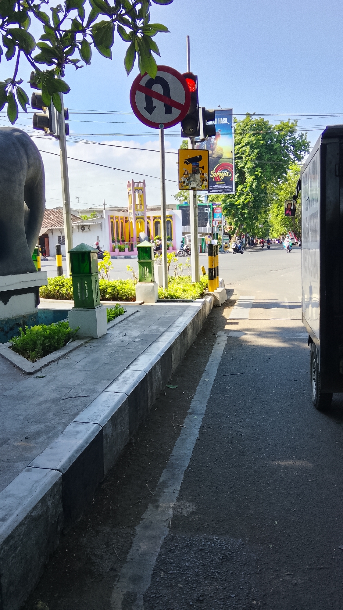 This image depicts a street scene in Pekauman, Indonesia, on a sunny day. A traffic light is visible, with a red light illuminated. A U-turn prohibition sign is also present. In the foreground, a concrete sidewalk runs alongside the road. Several green lampposts are placed along the sidewalk, and there is a statue on the left side of the frame, partially obscured by foliage. In the background, colorful buildings and trees can be seen. Several motorcycles are on the road, indicating traffic. The road surface is asphalt, and there is a white line marking a lane. A dark-colored truck is parked on the right side of the frame.