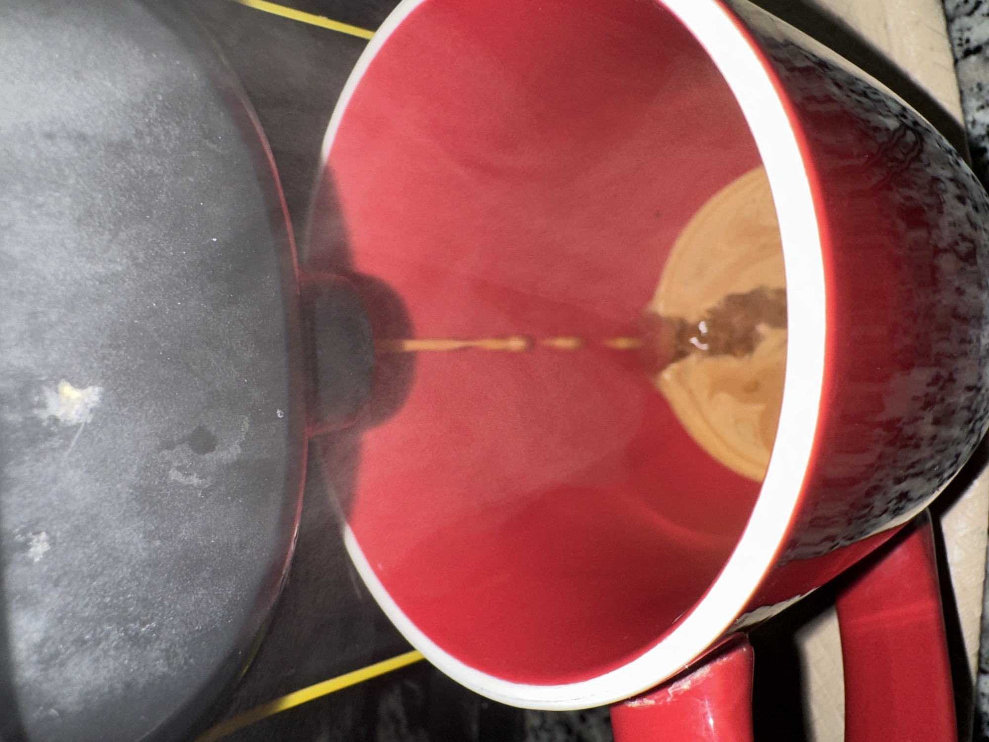 A red and white mug sits partially filled with tan liquid next to a gray pot. The mug is oriented with its open end toward the left, showing its red interior and the light tan liquid inside, which has darker swirling patterns. A thin stream of liquid connects the pot to the mug. The mug has a thick white rim and a visible red handle. The pot is gray and appears to be matte. The background shows a dark, possibly patterned surface underneath.