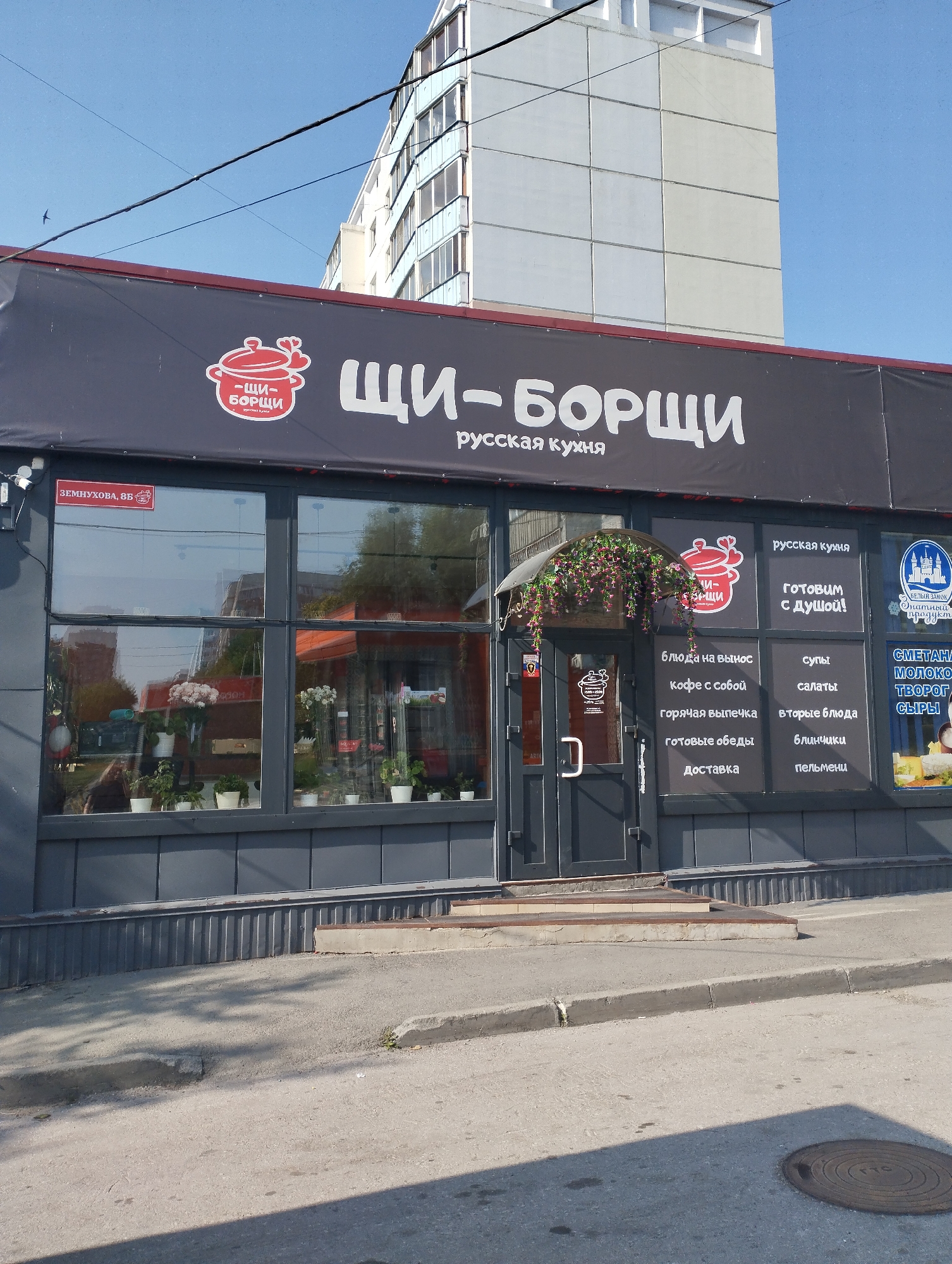The image shows the exterior of a restaurant named "ЩИ-БОРЩИ" (Shchi-Borschi), which specializes in Russian cuisine, located at Zemyukhova, 8B in Novosibirsk, Russia. The restaurant has a dark gray facade with a large sign featuring the restaurant's name in white Cyrillic letters and a red logo depicting a pot with hearts. Below the main sign, there is a smaller sign that says "русская кухня" (Russian cuisine).
The restaurant has large glass windows displaying various potted plants and flowers inside. The entrance is a dark gray door with a decorative archway adorned with artificial pink flowers. Several menu items are displayed on posters on the glass, including "блюда на вынос" (dishes to go), "кофе с собой" (coffee to go), "горячая выпечка" (hot pastries), "готовые обеды" (ready-made lunches), "доставка" (delivery), "супы" (soups), "салаты" (salads), "вторые блюда" (main courses), "блинчики" (pancakes), and "пельмени" (dumplings). A blue and white logo for "БЕЛЫЙ ЗАМОК" (White Castle), a brand of dairy products, is also visible.
The street in front of the restaurant is paved with asphalt and has a concrete curb. The building is situated in a city environment, with other buildings visible in the background, including a tall, modern building. The sky is clear and blue. There are no people visible in the image.