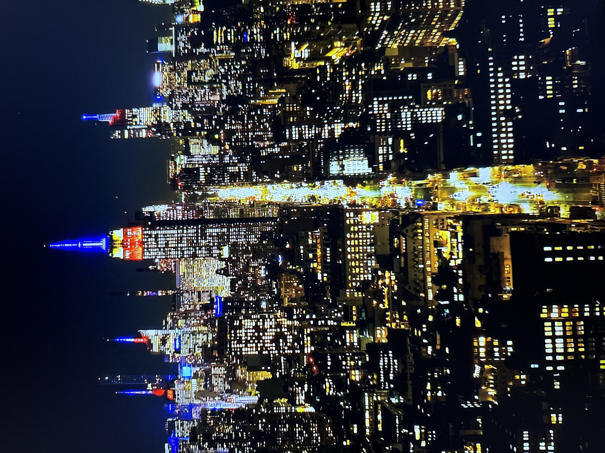 The image shows a nighttime aerial view of a densely populated cityscape with numerous illuminated buildings. The prominent structure in the center, illuminated with red and blue lights, is identifiable as the Empire State Building in New York City. A long, well-lit avenue, likely Fifth Avenue, runs through the heart of the scene, with visible traffic and streetlights. The surrounding buildings are a mix of skyscrapers and smaller structures, all displaying a multitude of glowing windows, indicative of a busy urban environment. The sky is dark, suggesting late evening or nighttime. The image does not depict any scenes from Burgas, Bulgaria.