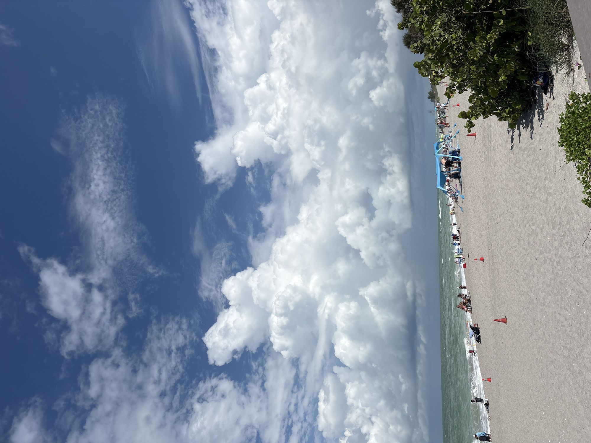 This image captures a sunny day at Englewood Beach in Englewood, Florida, United States. The sky is a vibrant blue, dotted with dramatic white cumulus clouds, some of which are quite large and puffy. The ocean water is a choppy, greenish-blue, with moderate waves rolling towards the shore. The beach itself is covered in light gray sand.
Several people are visible enjoying the beach. In the foreground, there are a few orange traffic cones placed on the sand. Further back, a blue canopy tent is set up, providing shade for a group of people. Other individuals are scattered across the beach, some sitting in chairs, others standing or walking. Some people are in the water. To the right of the image, lush green vegetation, including bushes and grasses, grows along the edge of the beach. The overall scene depicts a typical day at a populated beach with pleasant weather.