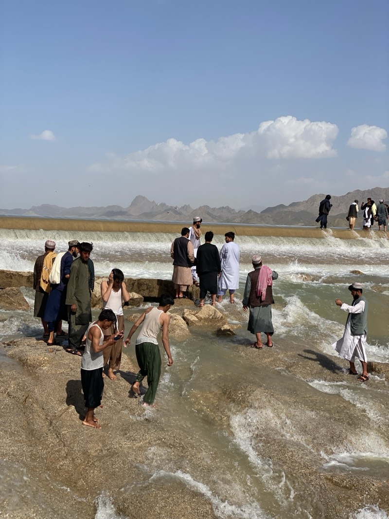 The image captures a scene at a river or waterway in Kim, Afghanistan, featuring a small dam or weir. Several men are present, some dressed in traditional Afghan clothing, including turbans and long shirts. Some of the men are wading in the water, while others stand on the dam structure. One individual appears to be using a mobile phone. The background consists of mountains and a blue sky with some clouds. The water is turbulent, with visible currents flowing over the dam. The terrain appears rocky and uneven.