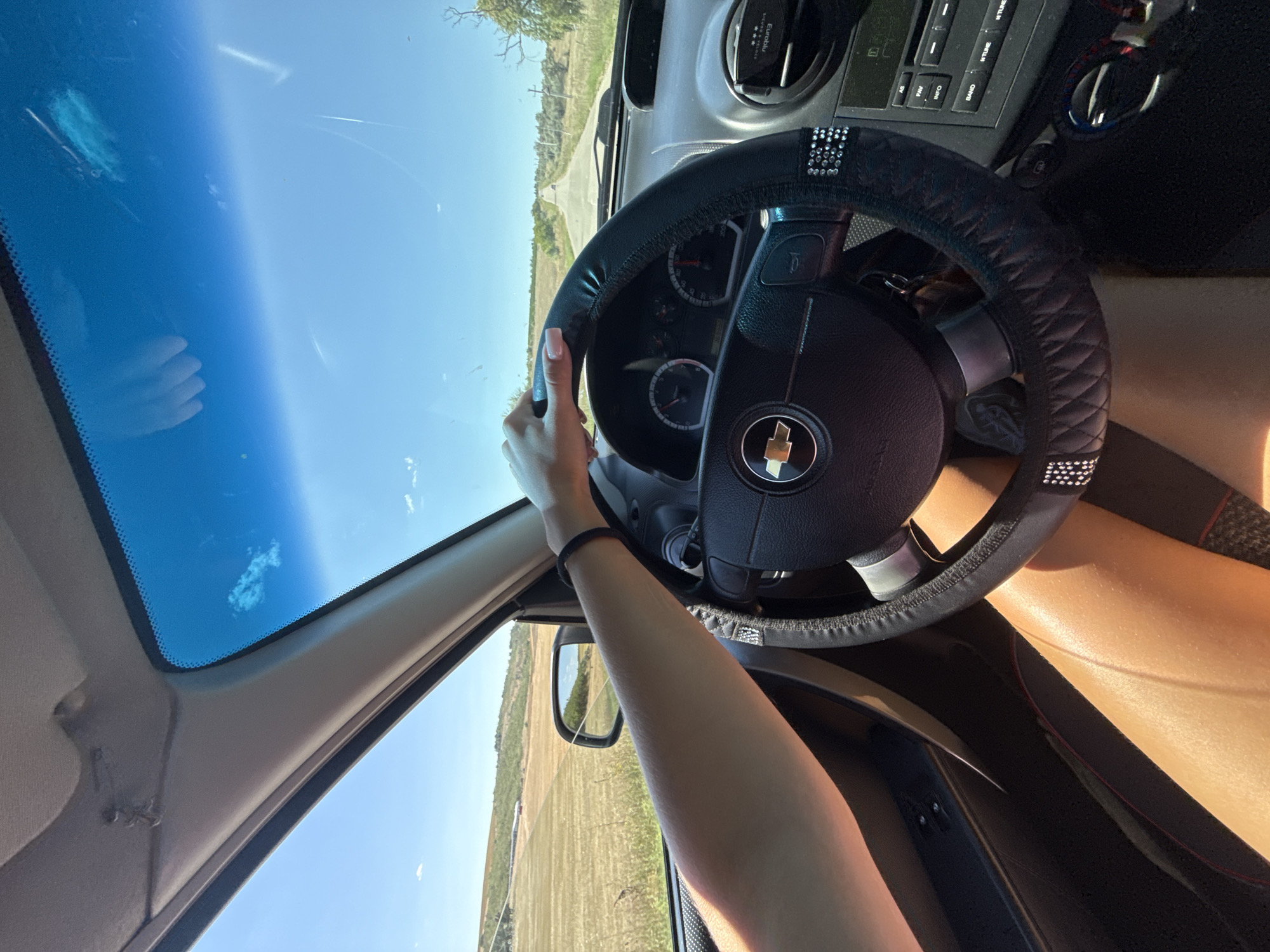 This is a first-person perspective image from inside a car driving on a rural road. The driver's left arm and hand are visible, resting on the steering wheel, which is covered in black faux leather with diamond accents. The dashboard is visible, including the steering wheel, instrument cluster, and part of the center console with a radio. Sunlight streams into the car, illuminating the driver's legs. Outside the car, a dry, grassy field stretches to the horizon, with a road receding into the distance under a clear blue sky with a few wispy clouds. The scene suggests a sunny day in a rural area. It is not possible to determine the exact location within Basarabeasca, Moldova, from this image alone.