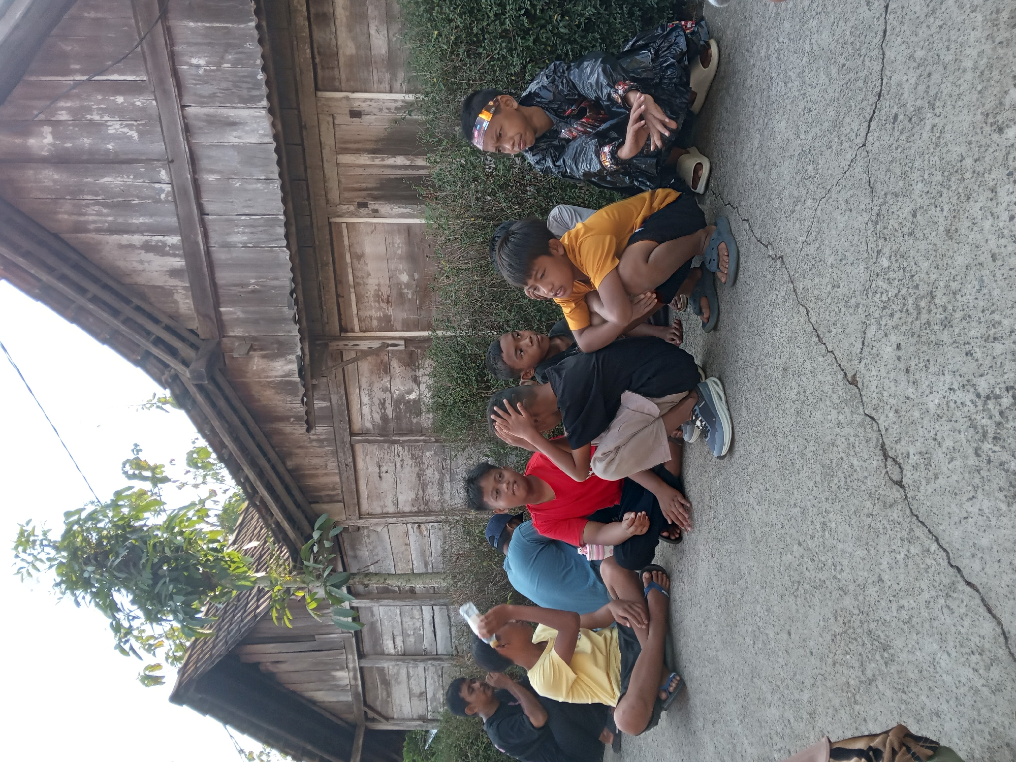 A group of Indonesian boys and a man are sitting on the ground outside a rustic wooden building in Kedunggudel, Indonesia. The building has a weathered, light-colored wooden exterior with vertical planks and a corrugated metal roof. A tall tree with green leaves grows in front of the building, and a dense hedge runs along the base.
In the foreground, seven individuals are visible. On the far left, a man in a black shirt and pants is partially obscured. Next to him, a boy in a yellow t-shirt and black shorts is holding up a plastic water bottle. To his right, a boy in a red t-shirt and black pants is looking to his left. In front of him, another boy in a black t-shirt and tan pants is crouched. Further to the right, a boy in an orange t-shirt and black shorts is looking away. Next to him, a boy is dressed in a black, shiny, possibly plastic outfit with colorful accents and a headband, and he is looking to his right. Another boy in a blue shirt and a blue cap is visible in the background, partially behind the other seated individuals. The ground is concrete with visible cracks.