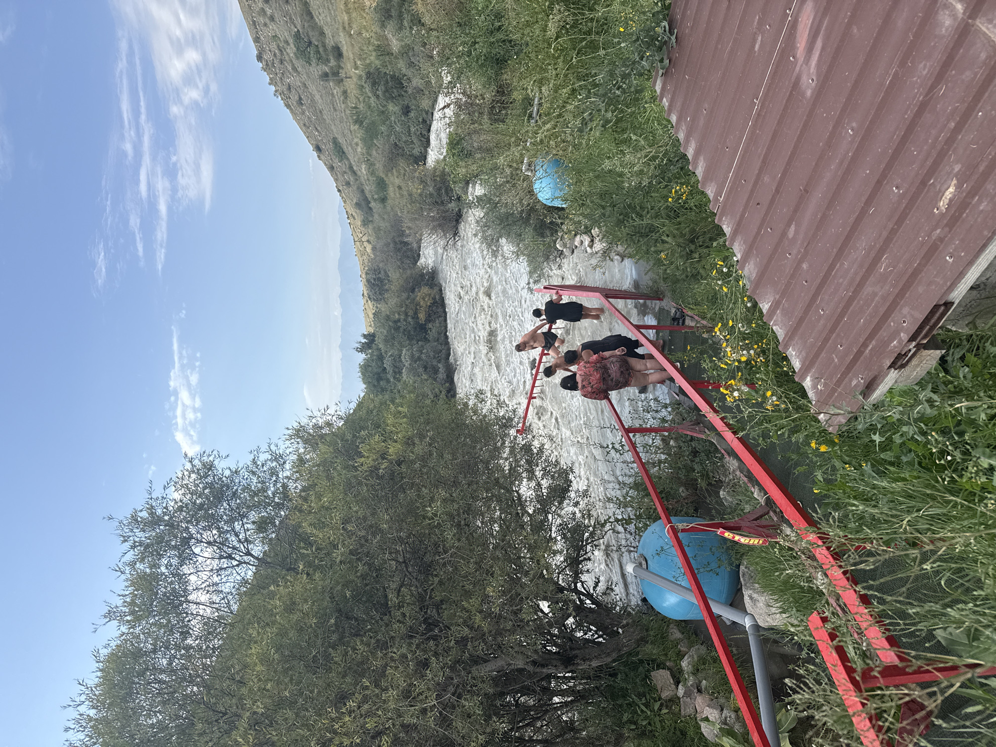This is a wide shot of a natural river with several people interacting with the water. In the foreground, a red railing leads down to the river. A few individuals, appearing to be tourists or locals, are in the water or on the path approaching it. A large blue tank is visible to the left, near the railing. The river itself is fast-moving and appears to be a popular spot for recreational activities. The background is characterized by lush greenery and rolling hills under a bright blue sky with scattered clouds. The scene evokes a sense of natural beauty and outdoor recreation. The location is identified as the city of Ak-Suu, Kyrgyzstan.