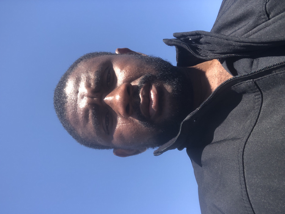 The image displays a close-up, low-angle portrait of an adult male. The individual has dark skin, short dark hair, and a full beard. His face shows a noticeable sheen, consistent with perspiration or direct sunlight. He is wearing a dark-colored, zippered jacket with a raised collar, the zipper partially open. The background is an expanse of clear, bright blue sky, indicating an outdoor daytime setting. Strong direct sunlight illuminates the subject, causing highlights on his skin and casting shadows beneath his chin and on his neck. The scene is located in the city of Unknown, Saudi Arabia.