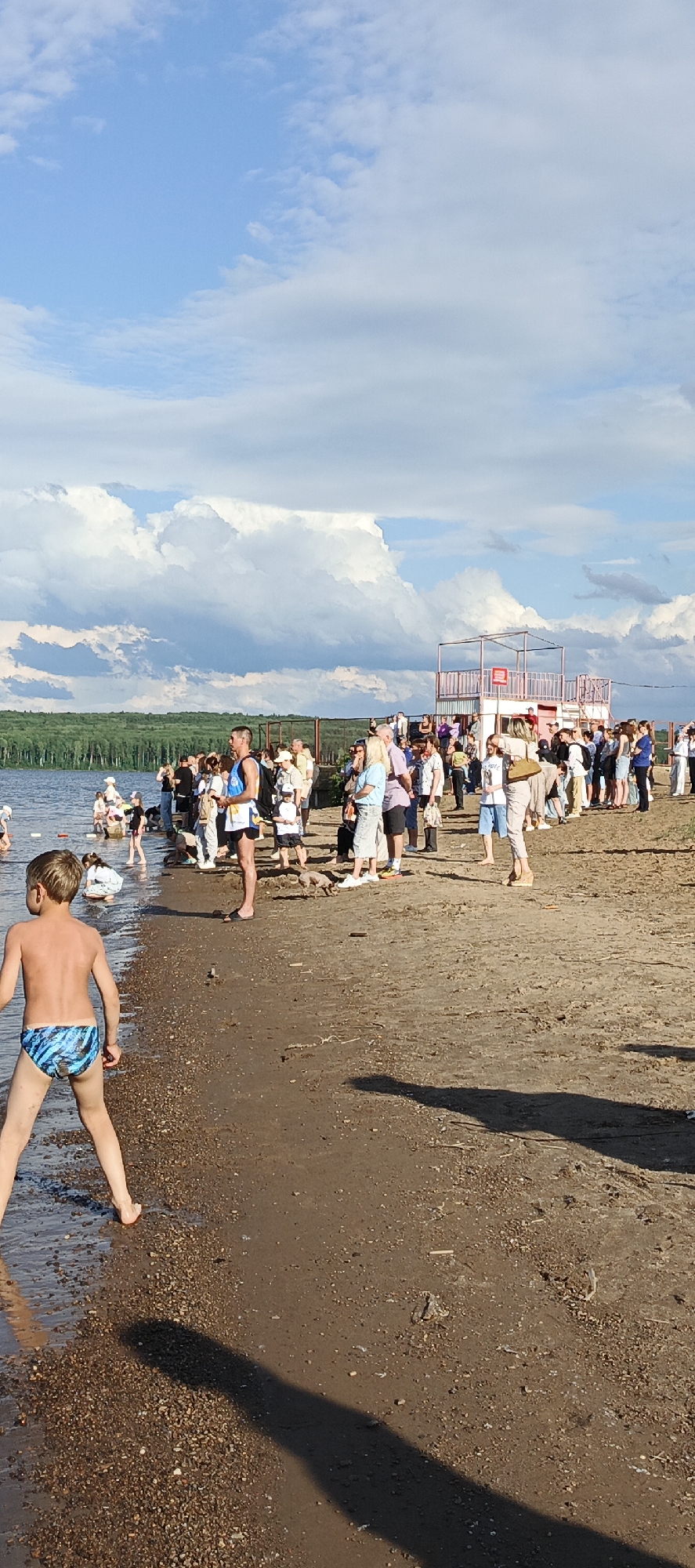 A crowded sandy beach runs along a body of water, with a dense green forest visible across the water. The sky is partly cloudy with large white cumulus clouds. In the foreground, a young boy in blue patterned swim trunks stands on the wet sand, facing away from the camera. To his left, the water laps at the shore. Further back, a group of people are gathered near the water's edge. In the middle ground, a group of people stand on the beach, some observing the water, others talking. A small, elevated, open-air structure with a pink railing and a roof stands on the right side of the beach, and more people are gathered around and near it. The beach is composed of sand and small pebbles, with some debris scattered on the ground. The lighting suggests it is late afternoon or early evening. The location is the beach area of the Kama River embankment in Chaikovsky, Russia.