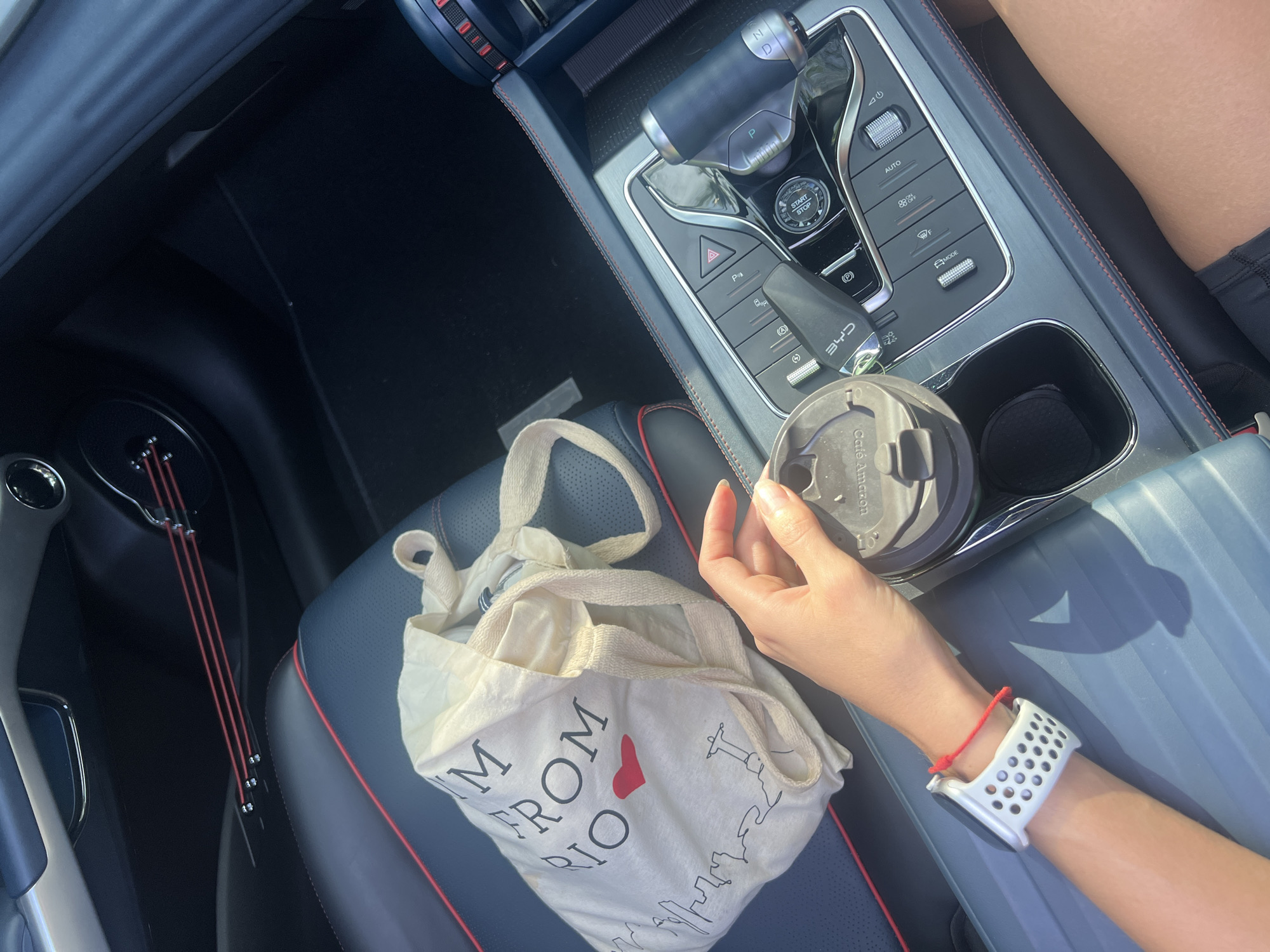 A person's right arm, adorned with a white smartwatch and a red string bracelet, rests on a car's console next to a closed, dark coffee mug, placed in a cupholder. A white tote bag with the text "I'M FROM RIO" sits on the car seat next to the console. The gear shift and console controls of the vehicle are visible above the cup holder, and a black car mat can be seen in the left foreground. The environment indicates the inside of a car on a sunny day.