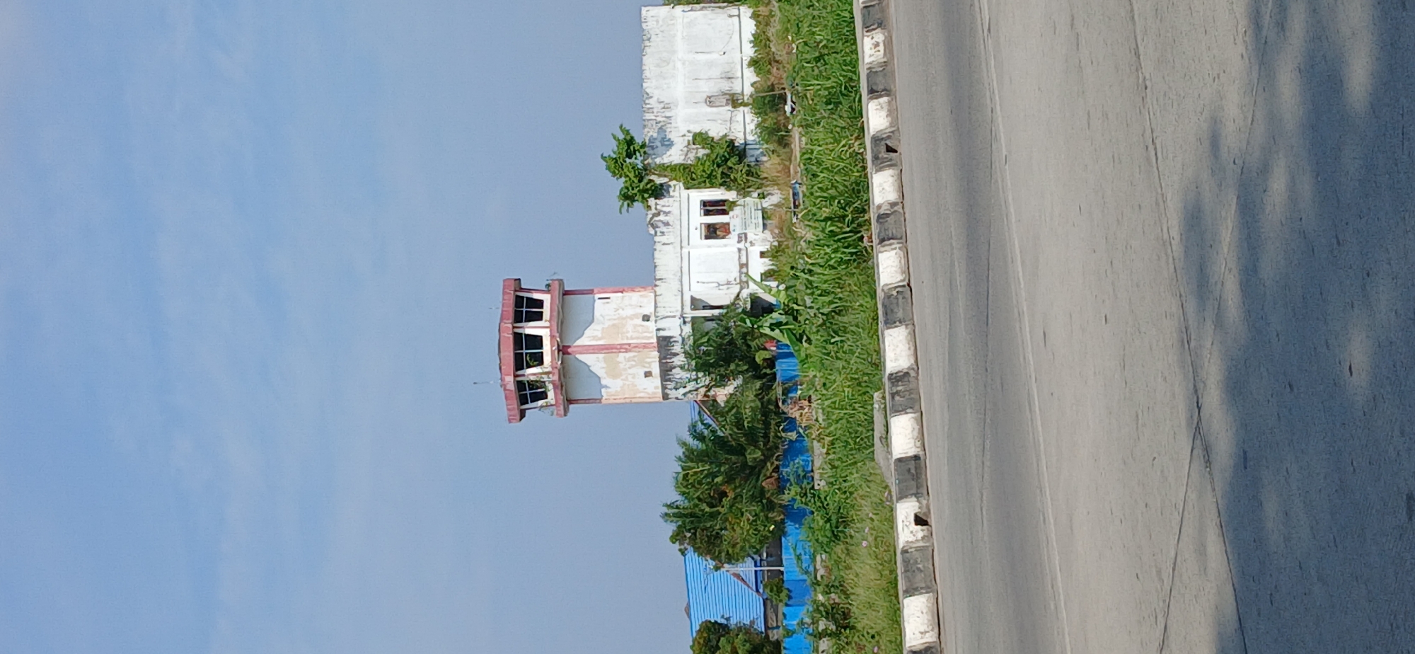 The image shows an air traffic control tower and an adjacent building in Samarinda, Indonesia. The tower is octagonal with a red trim around the top and a white body. It has large windows that are divided into panes. The building next to it is white and appears to be in disrepair, with peeling paint and overgrown vegetation. A blue roofed structure is visible to the left of the tower. The scene is framed by a road in the foreground with a concrete barrier. Lush green grass and trees surround the buildings. The sky is clear and blue.