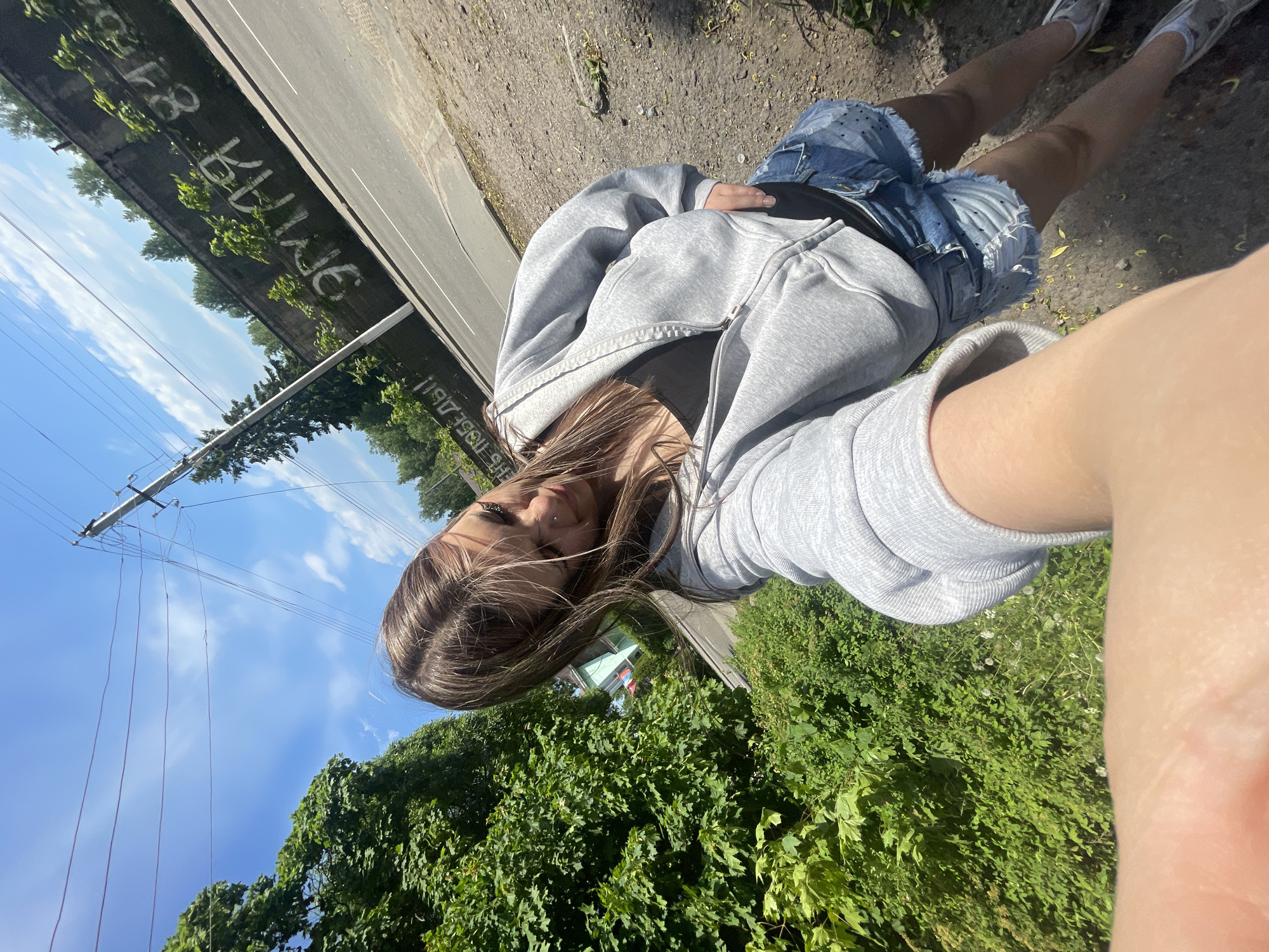 The image depicts an outdoor, high-angle selfie of a young woman taken on a sunny day in Priozersk, Russia. The woman, positioned in the lower center, has long, light brown hair, a nose piercing, and is wearing a light gray zipped hoodie over a dark top and ripped blue denim shorts with small dark dots. Her left arm, holding the camera, is visible in the foreground. She is wearing light-colored, possibly gray, closed-toe shoes.
The background features lush green foliage from large trees and bushes on the left. To the right, an asphalt road runs parallel to a tall, weathered concrete wall. The wall displays white graffiti, including "ЭТО ПОБЕДЫ!" (This is Victory!) partially visible on the lower left, and "ЭТОТ Я 81" (This one I 81) visible on the upper right. A utility pole with multiple power lines stands between the trees and the wall on the right. The ground in the foreground is a rough mix of dirt and small stones, with some green growth and scattered debris, including what appears to be discarded plastic items. The sky above is clear blue with scattered white clouds. The overall scene suggests an urban or semi-urban environment with significant natural elements.
