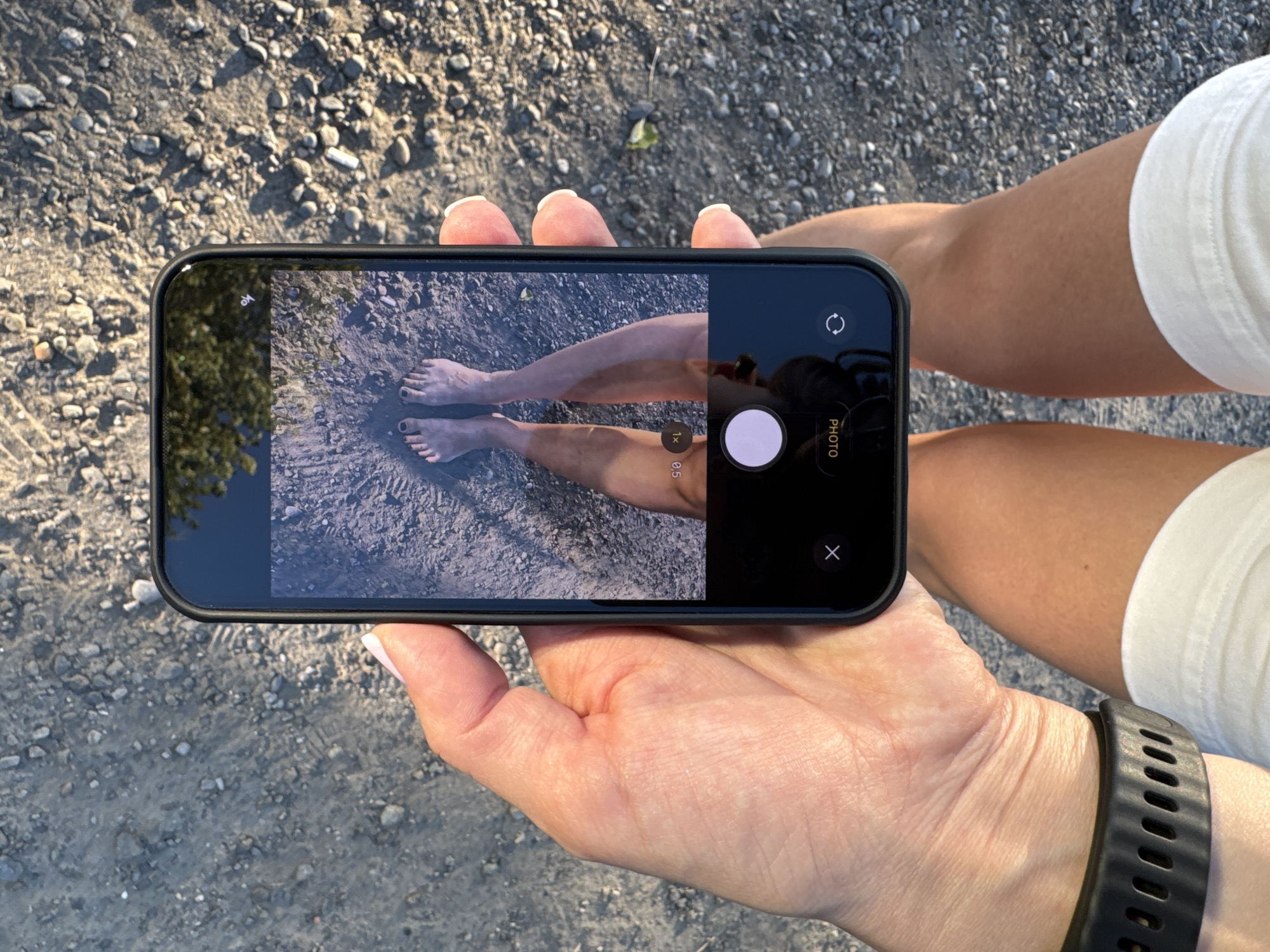 A person's hands hold a smartphone, displaying a close-up shot of their feet on a gravelly surface. The phone screen shows the camera interface, with options like "PHOTO" and a zoom level indicator. The person is wearing white shorts and a black smartwatch. The background is composed of gravel and a hint of greenery at the bottom. The image is taken outdoors during daylight. The scene does not depict any specific landmarks or identifiable features of Sochi, Russia.