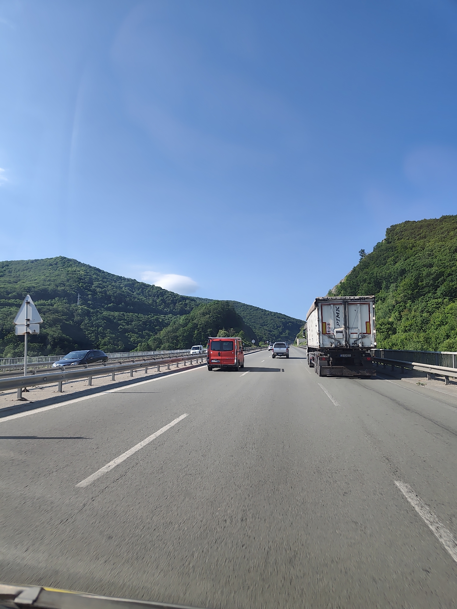 A clear, bright day with a bright blue sky is depicted, with a few wispy clouds visible. The scene is a highway passing through a mountainous, heavily forested region. The highway has multiple lanes and is bordered by guardrails on either side. Several vehicles are on the road, including a white semi-trailer truck with "P&K" branding on its trailer, a red van, and a dark blue car. The surrounding landscape is dominated by lush green mountains covered in dense trees, with some rocky outcrops visible. The perspective suggests the image was taken from inside a moving vehicle. The location is identified as being on the highway near the village of Malo Buchino, Bulgaria.