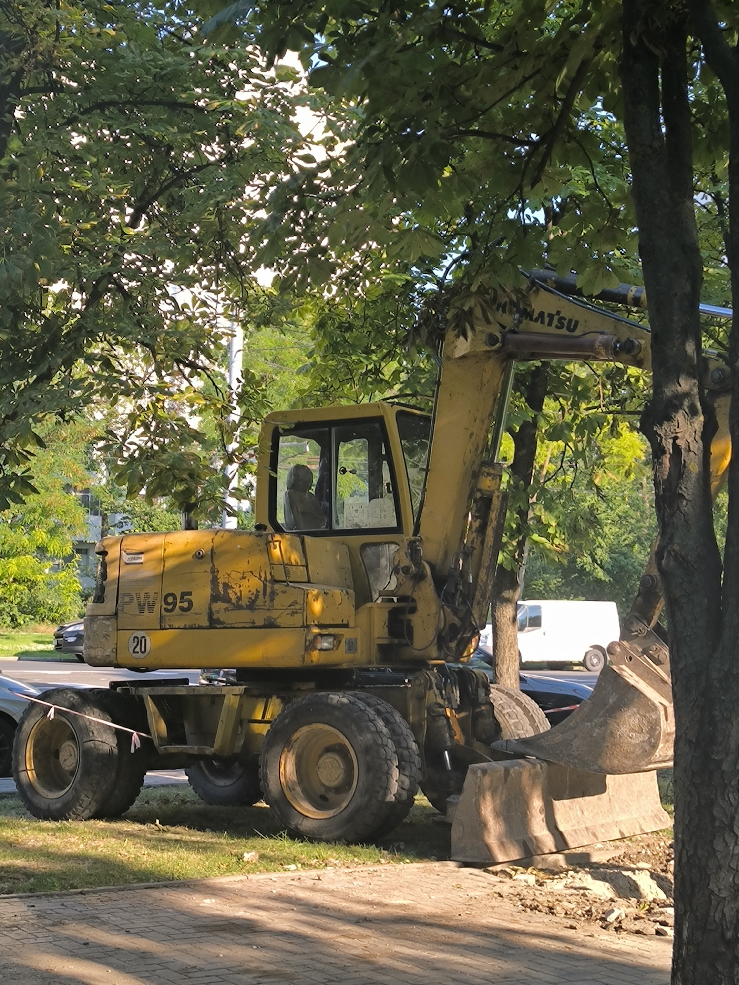 An outdoor, medium shot captures a yellow Komatsu PW95 wheeled excavator parked on a sidewalk next to a street in Chișinău, Moldova. The excavator is positioned beneath the dappled sunlight filtering through the dense green foliage of mature trees lining the street. The machine's prominent yellow paint is weathered and shows signs of use. Its large bucket is lowered, resting on a large concrete barrier which appears to be part of road construction or maintenance. A reflective marker, likely a speed limit sign, shows the number "20" on the side of the excavator's front. In the background, two vehicles are visible: a dark-colored car on the street and a white van parked further down. The sidewalk is paved with grey concrete slabs, and a grassy strip with scattered debris runs alongside it. The overall scene suggests ongoing public works or construction activity in a residential or commercial area of the city.
