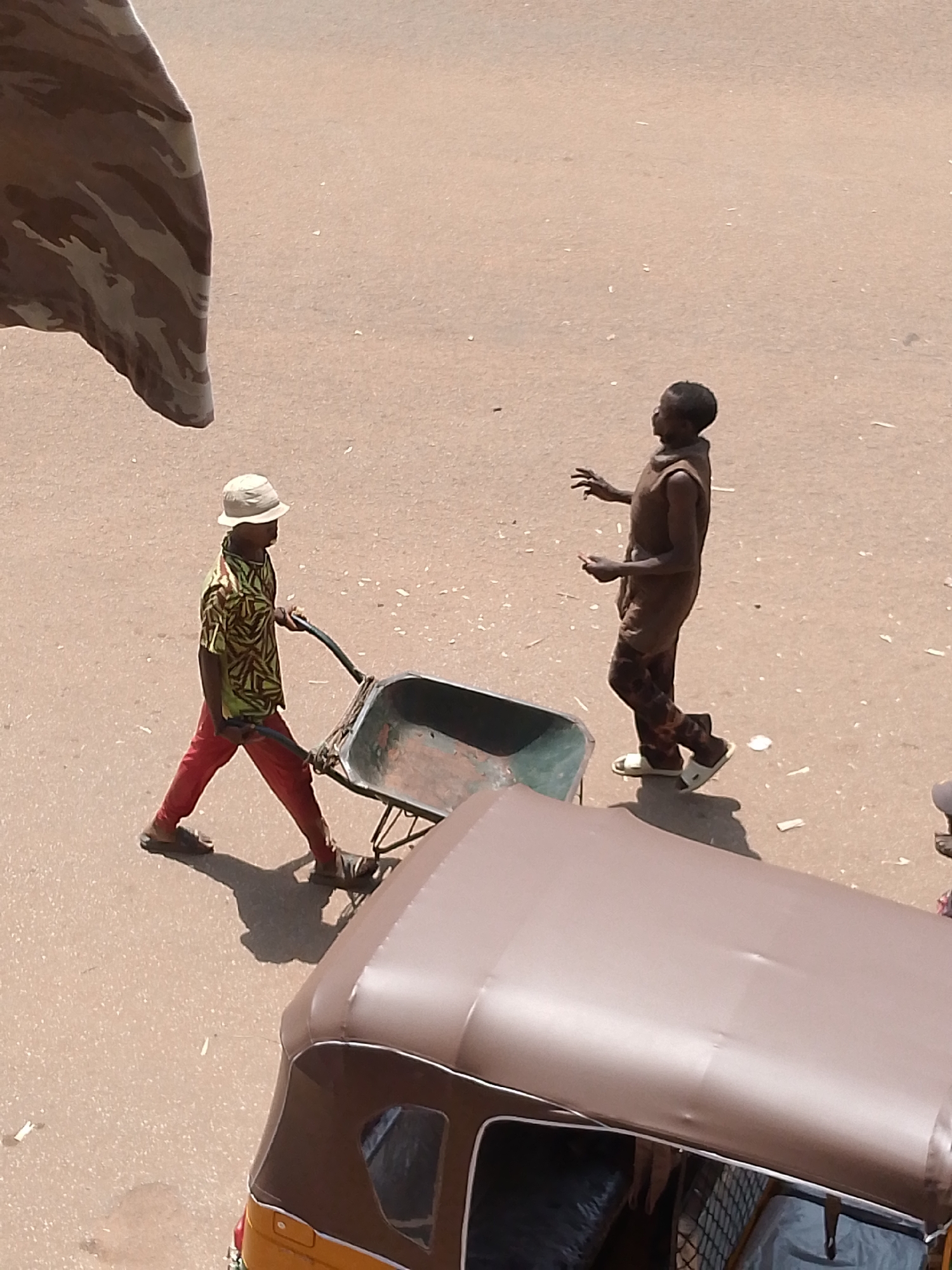 In Bukuru, Nigeria, an overhead shot captures a dusty, paved street scene. Two boys are the primary subjects. On the left, a boy wearing a light-colored bucket hat, a brightly patterned short-sleeved shirt in yellow and green, and red pants pushes a large, empty wheelbarrow. He is wearing dark sandals. To his right, a second boy, dressed in a brown sleeveless tunic and patterned brown pants, stands with his hands outstretched, appearing to be in the motion of catching or playing with something unseen. He is wearing white sandals. In the foreground, the brown canopy of a three-wheeled auto-rickshaw partially obscures the lower portion of the image, its windshield visible. The ground is a pale, sandy color with scattered debris. A camouflage-patterned fabric hangs from the top left corner of the frame, also partially obscuring the scene.