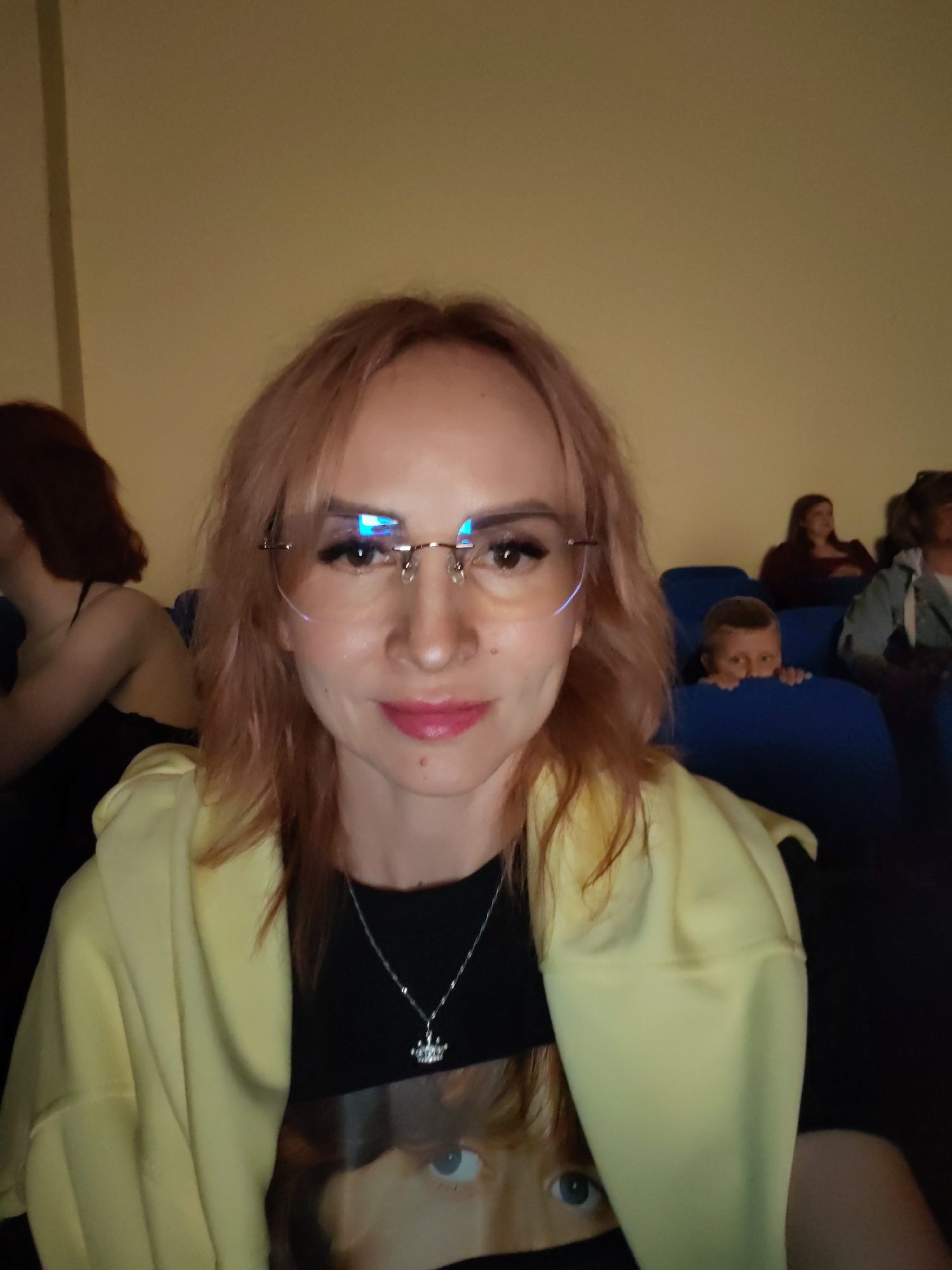A woman with light-colored hair and glasses is looking at the camera, smiling. She is wearing a yellow hooded jacket over a black t-shirt with a picture of a child on it. She is also wearing a silver necklace with a crown pendant. In the background, other people are seated in a dimly lit room, possibly a movie theater or lecture hall. A child is peeking over a seat in the background. The environment appears to be indoors, with yellow walls and blue seats. The image is a selfie, taken from a low angle. The woman is in the foreground, while the other people and the environment are in the background. The location is not specified as being in the city of Chaikovsky, Russia.