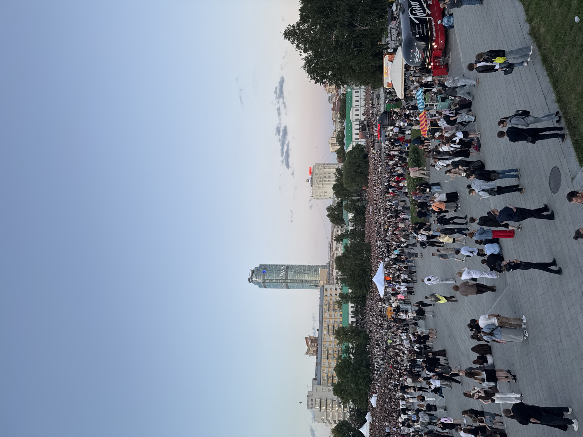 This is an outdoor, eye-level, wide-angle shot of a large public gathering in Yekaterinburg, Russia, likely on a warm evening given the light blue sky with scattered clouds and the attire of the people. The scene is dominated by a vast crowd filling a paved plaza in the foreground and midground, with a dense mass of people extending towards the background. Numerous individuals are visible, many standing and milling about, while some are seated. A few white tents are interspersed within the crowd.
In the background, the Yekaterinburg City Administration building and the prominent skyscraper Iset Tower are visible, along with other urban architecture and trees. A red food truck with "TORTO" branding is parked on the right side of the frame, and a smaller white food stall is nearby. The plaza is paved with grey interlocking stones, and some green grass is visible in the lower right corner. The overall impression is of a lively event or festival taking place in a city square.