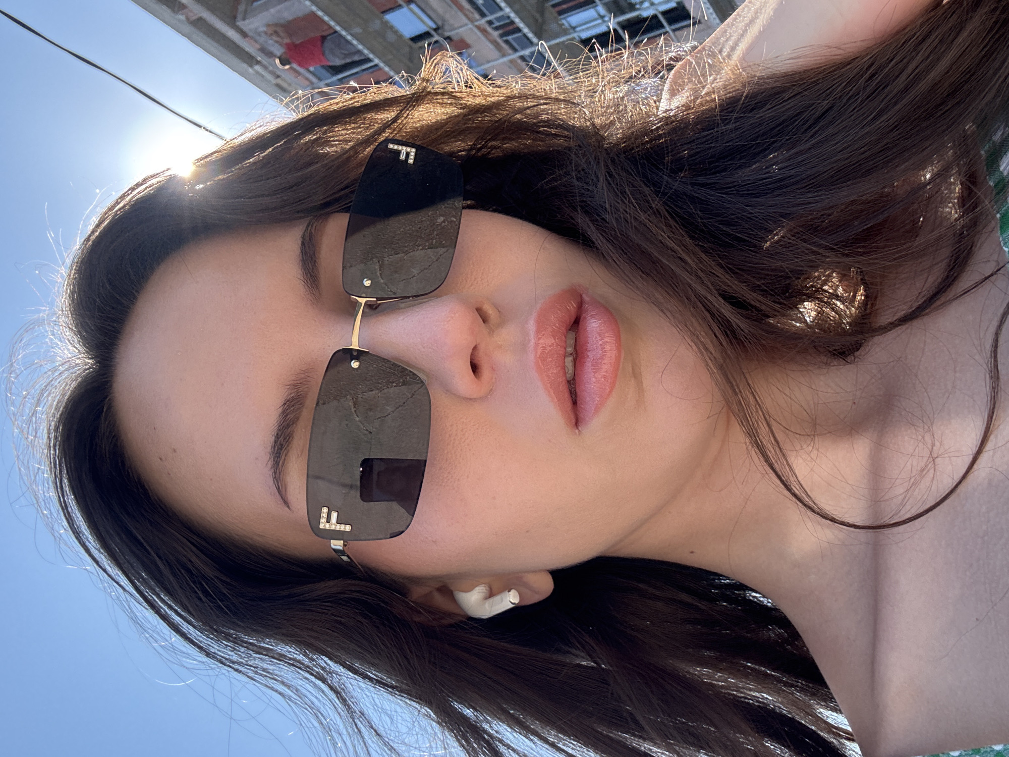 A medium shot captures a young woman with long, dark hair wearing sunglasses and white wireless earbuds. She is looking into the camera, and the sun is shining brightly, creating lens flare. In the background, an unfinished building with scaffolding is visible, and a person in a red shirt is working on it. The sky is clear and blue.