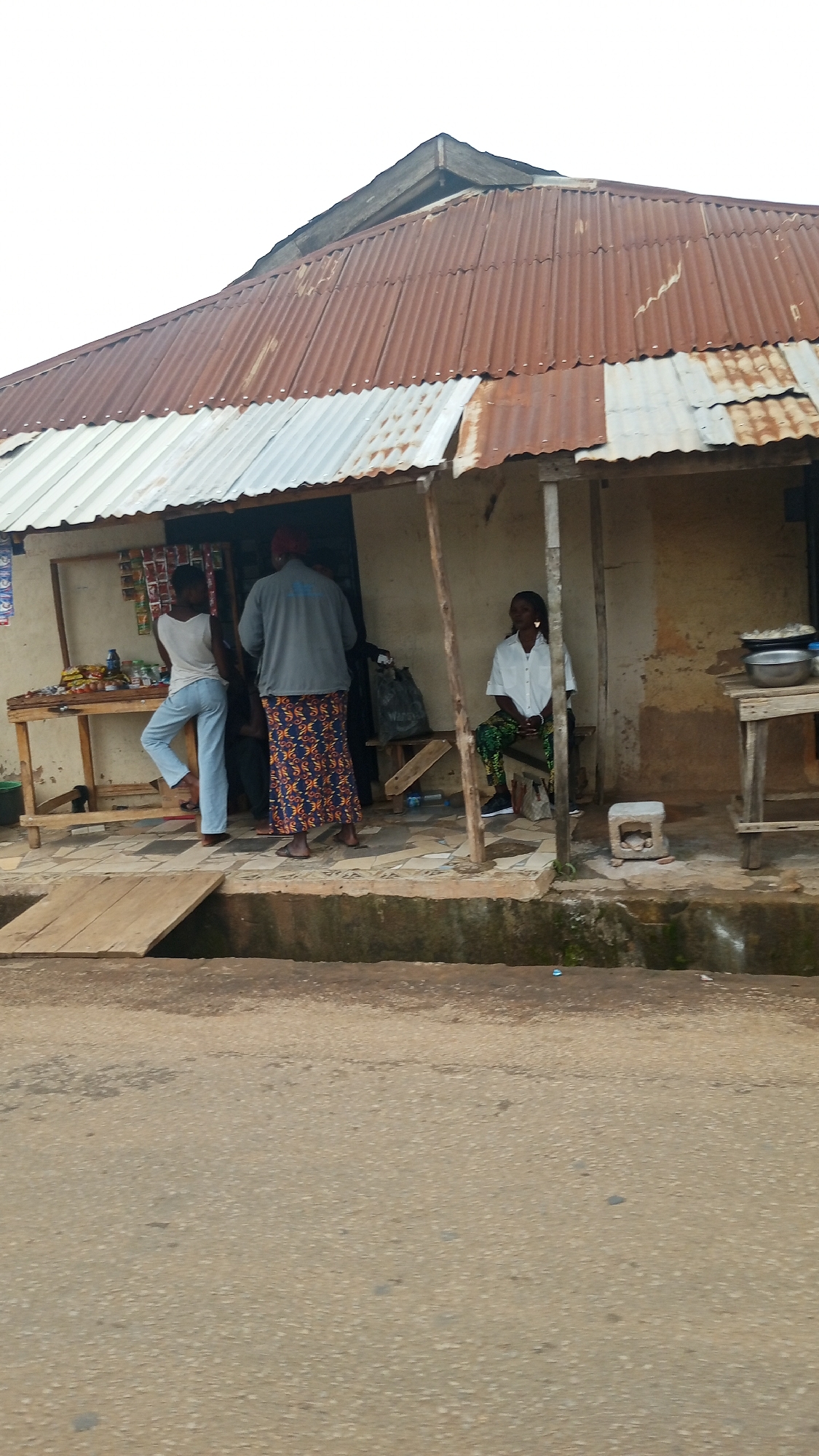 The image shows a street-level view of a small, open-air shop with a corrugated metal roof in Bukuru, Nigeria. Three individuals are visible. On the left, a young woman in a white t-shirt and light blue jeans stands near a wooden stall displaying various items, possibly food or merchandise. In the center, a person wearing a grey shirt and a colorful, patterned skirt stands with their back to the camera, seemingly interacting with the items on the stall. To the right, a woman in a white shirt and patterned green trousers sits on a bench. A small concrete block is placed on the ground near her. The shop itself is constructed with rough, earthy-colored walls. The foreground shows a paved road and a wooden plank laid across a drain. The overall scene depicts a typical informal marketplace setting.