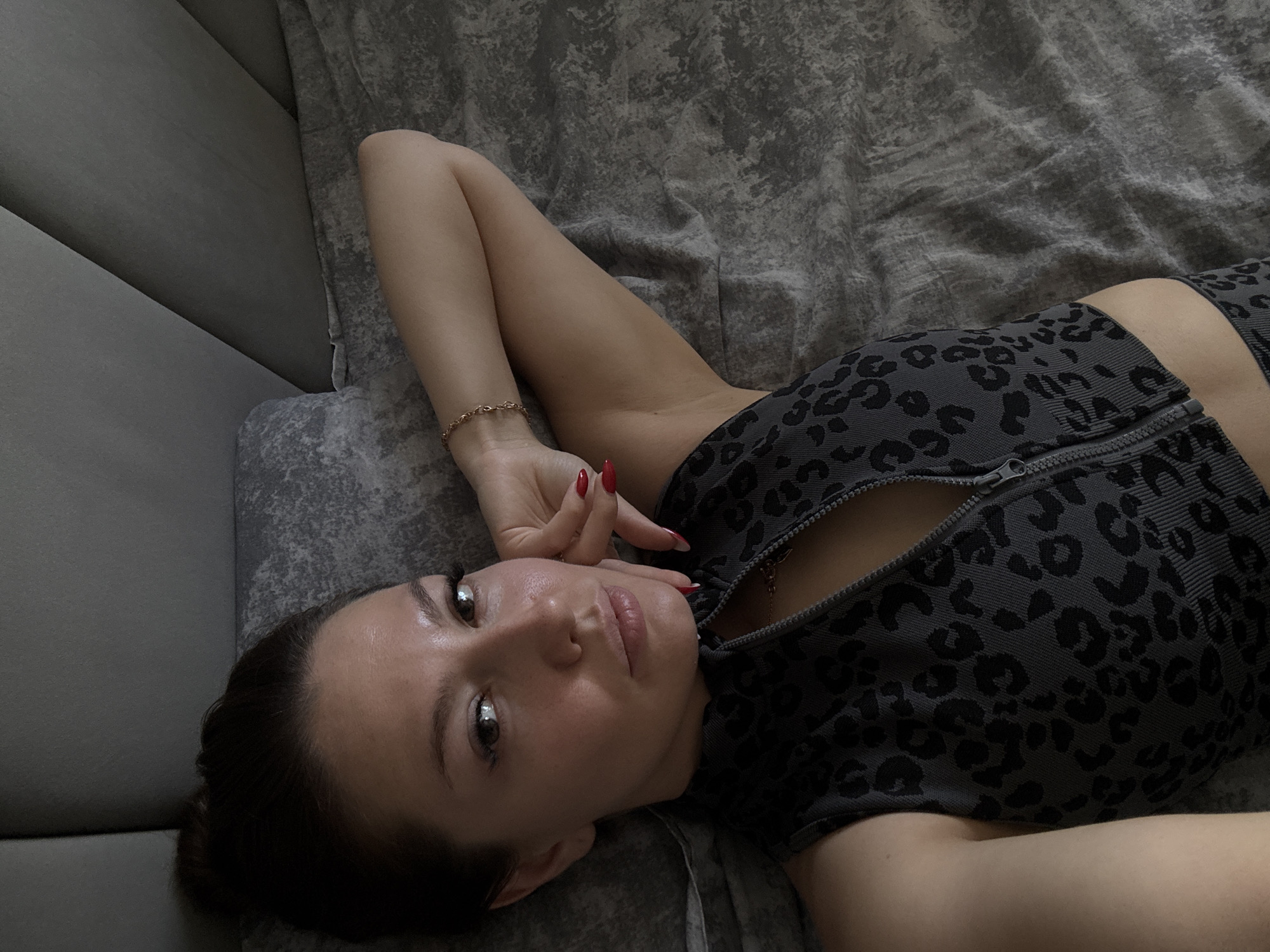 This media file is a close-up, overhead shot of a young woman lying on a textured gray surface, possibly a bed or sofa. She is looking directly at the camera with a relaxed expression. Her head is propped on her arm, which is bent at the elbow and resting behind her head. She has dark hair and appears to be wearing a grey top with a black leopard print pattern. The top has a zipper that is partially open, revealing a hint of her chest and a delicate gold necklace. Her fingernails are painted a bright red. The lighting is soft, suggesting an indoor setting with diffused light, possibly during the day. The background is a textured gray fabric, with a slightly lighter gray angled surface visible to the left, creating a sense of depth. The overall mood is intimate and casual.